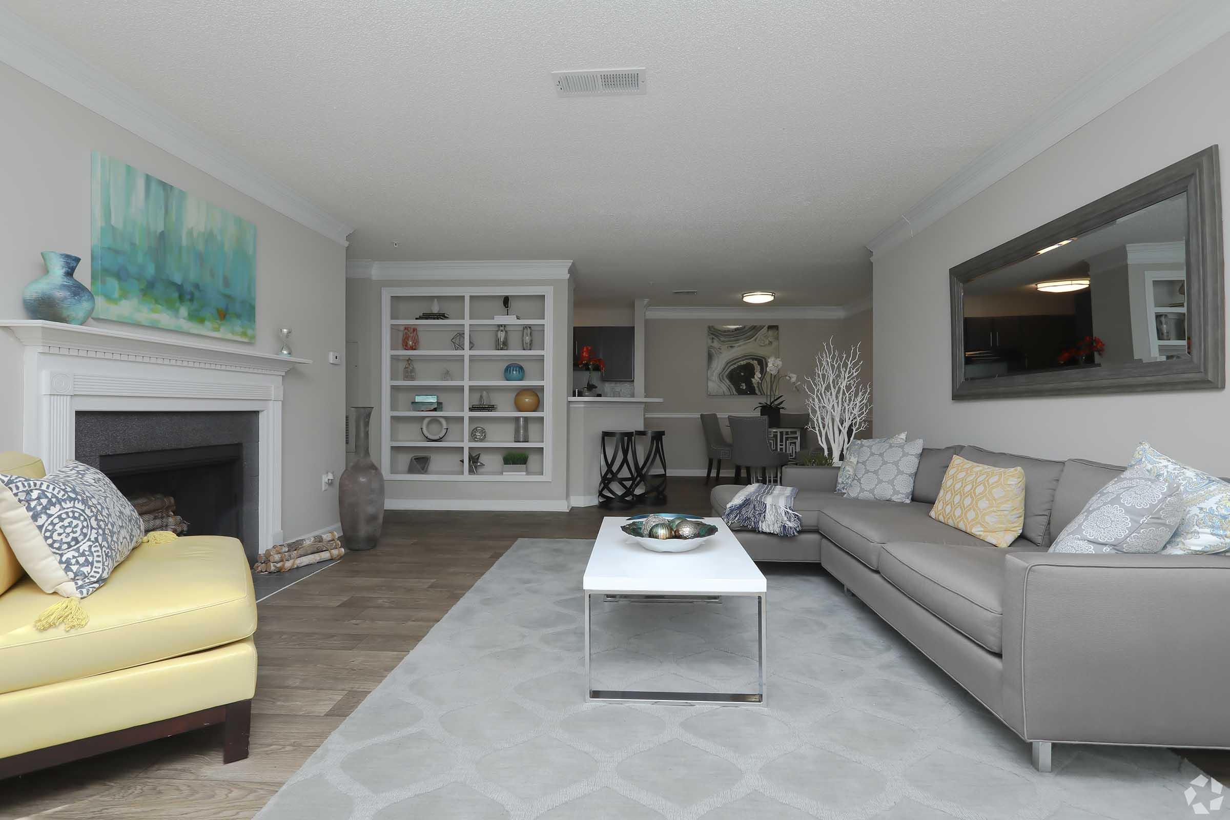 A modern living room featuring a light gray sofa with colorful throw pillows, a yellow accent chair, and a white coffee table with decorative items. A textured area rug covers the floor, and a fireplace is visible on the left. The room is well-lit, with a neutral color palette and decorative elements on shelves in the background.