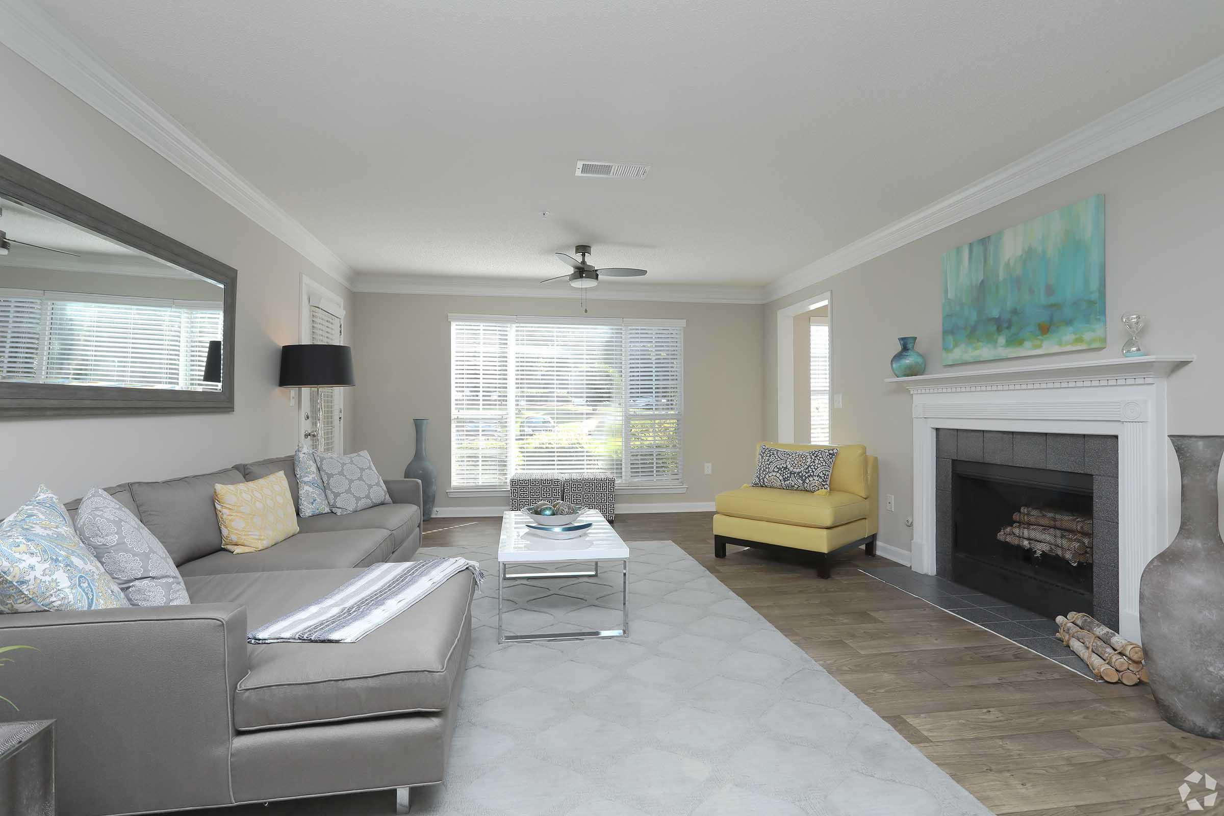 A cozy living room with a grey sectional sofa and a yellow accent chair. A white coffee table sits on a grey rug, and a modern fireplace is to the right. Large windows provide natural light, and decorative items, including a vase and artwork, add a touch of style to the space.
