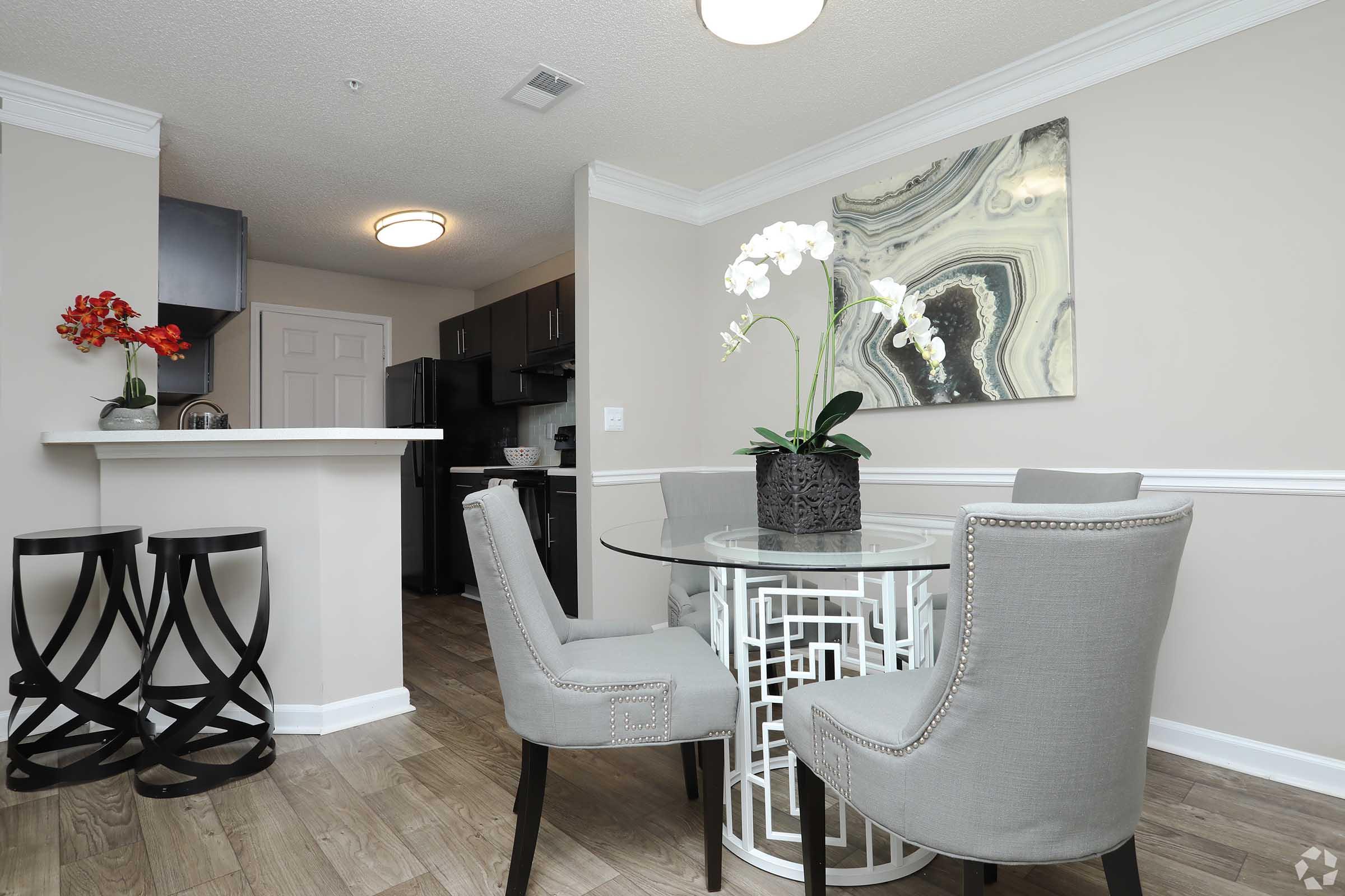 A modern dining area featuring a round glass table with stylish gray chairs. On the table, there is a decorative pot with white orchids. In the background, there is a small kitchen area with dark cabinetry, and a piece of abstract wall art hangs above the table. Bright and contemporary decor enhances the ambiance.