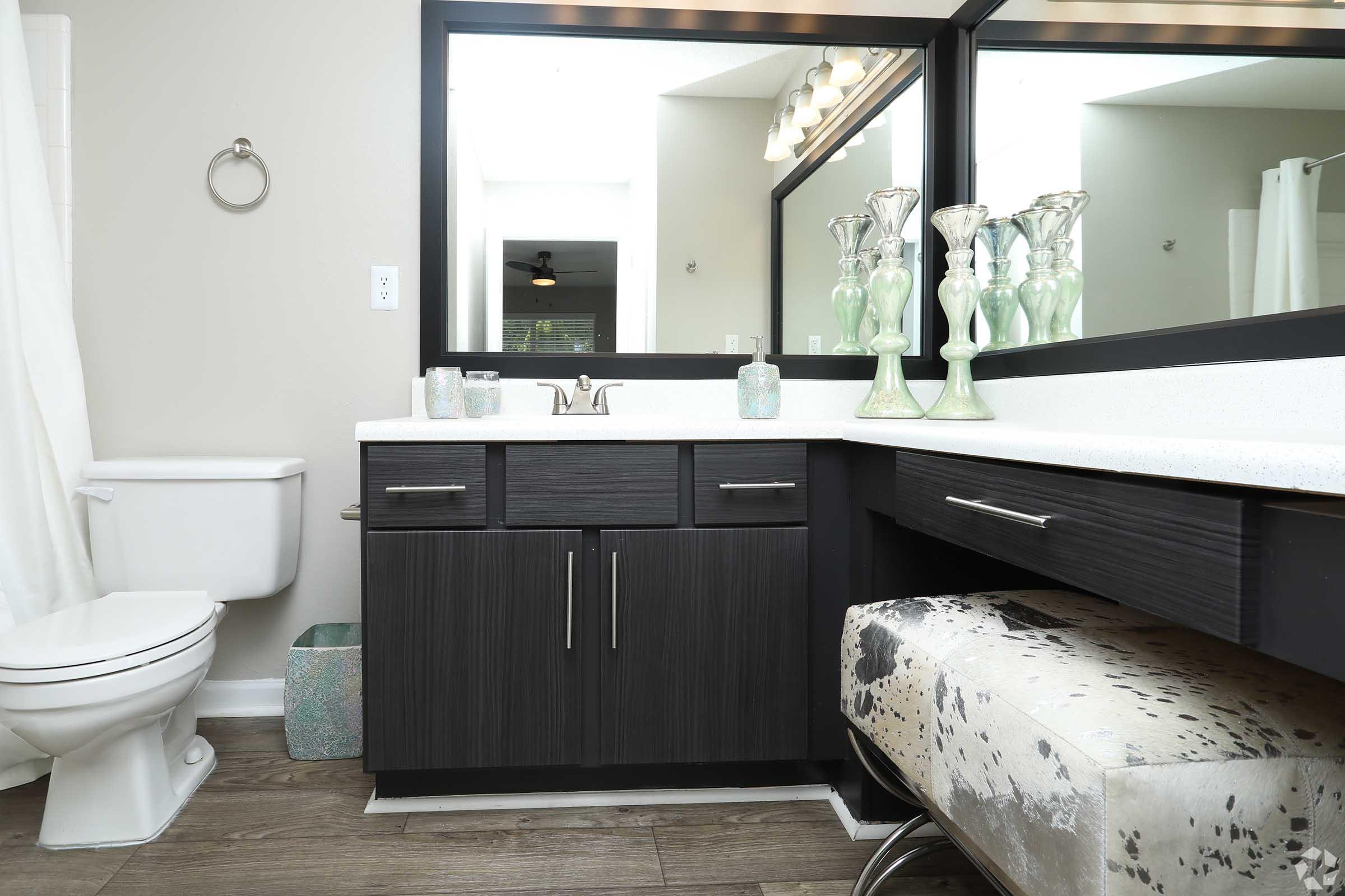 A modern bathroom featuring a stylish vanity with a white countertop, dark cabinetry, and a large mirror. Decorative bottles are displayed on the countertop, alongside a plush upholstered stool. A white toilet is visible to the left, and natural light filters in from a window above.