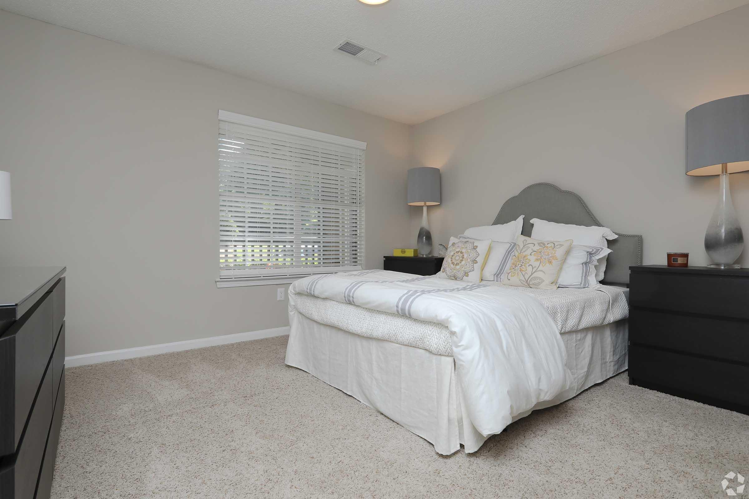 A cozy, well-lit bedroom featuring a queen-sized bed with decorative pillows and a light-colored bedspread. The room has two lamps on bedside tables, a dark wood dresser, and large windows with white blinds, creating a serene atmosphere with neutral-colored walls and plush carpeting.