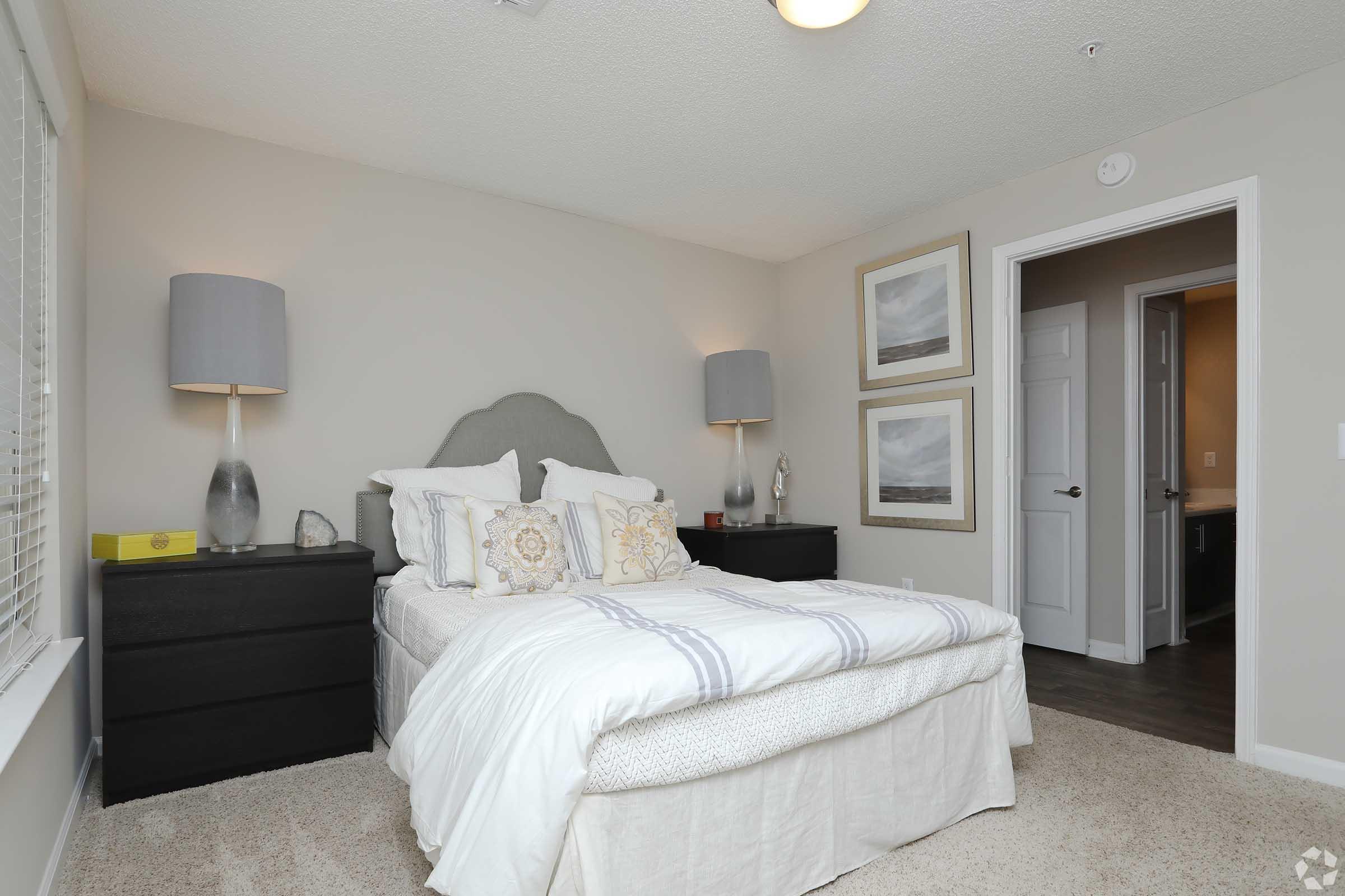 A modern bedroom featuring a neatly made bed with decorative pillows, bedside lamps on dark wood nightstands, and two framed artworks on the wall. The room has light gray walls and soft carpet, creating a cozy atmosphere. An open door leads to another room in the background.