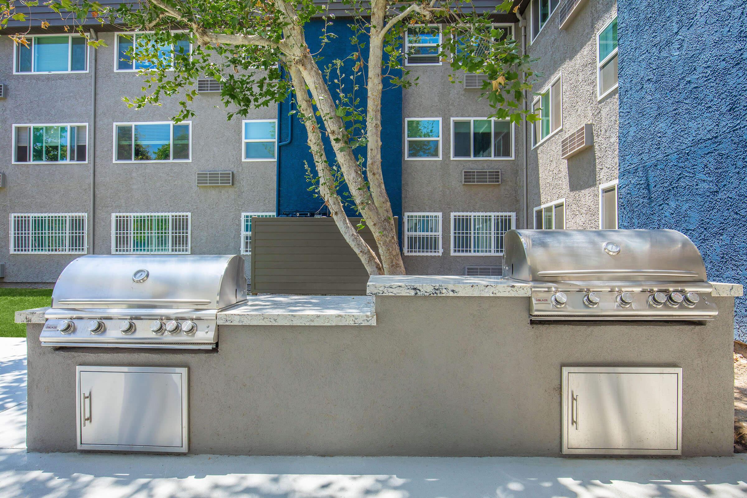 Outdoor barbecue area featuring two stainless steel grills with a stone countertop, surrounded by green grass and residential apartment buildings in the background. A large tree provides shade, and the building façades are painted in gray and blue tones.