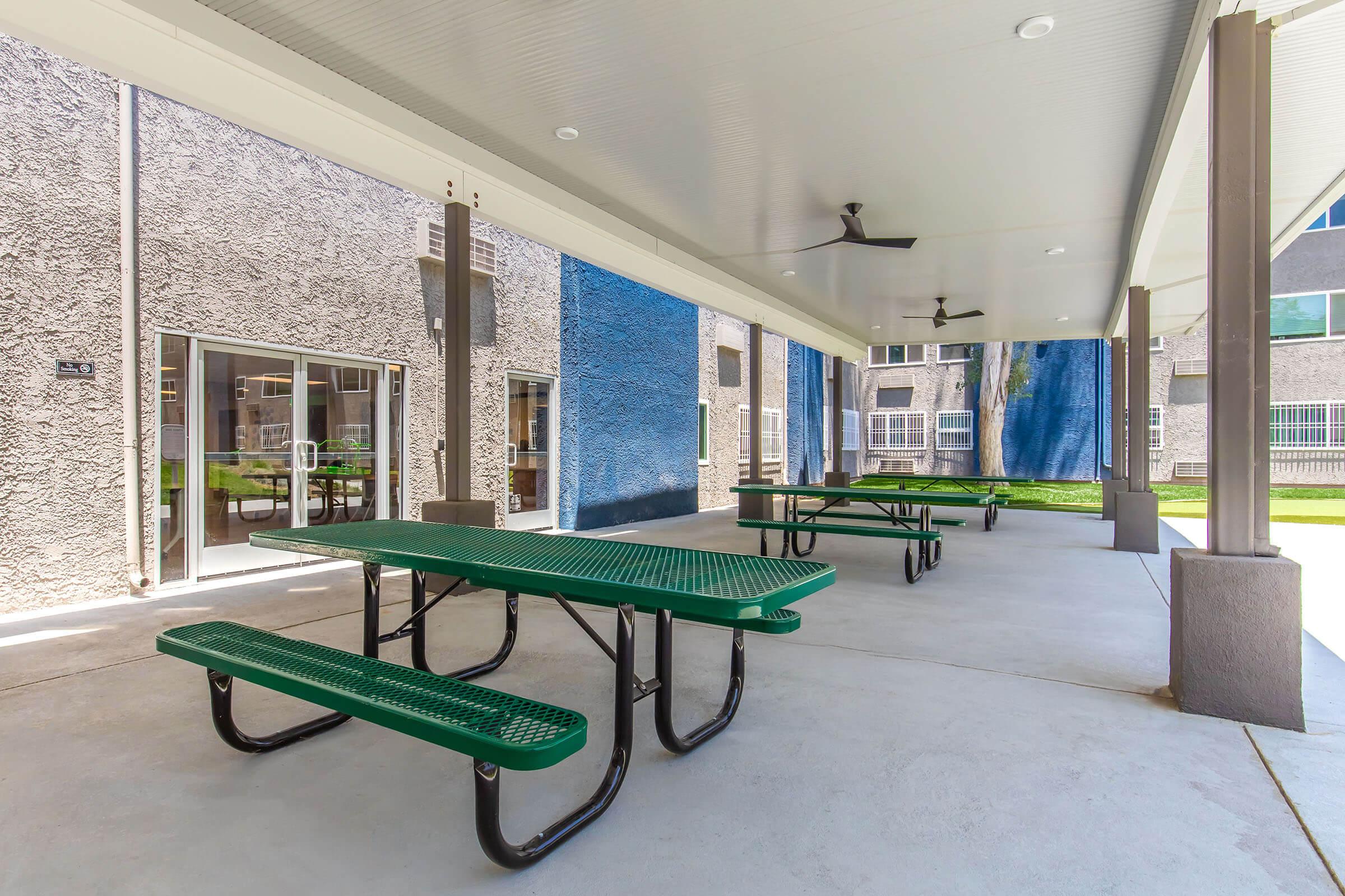 Covered outdoor gathering area with green picnic tables and ceiling fans, featuring a concrete floor and walls painted in gray and blue. The space is well-lit and surrounded by windows, providing a comfortable setting for social activities.