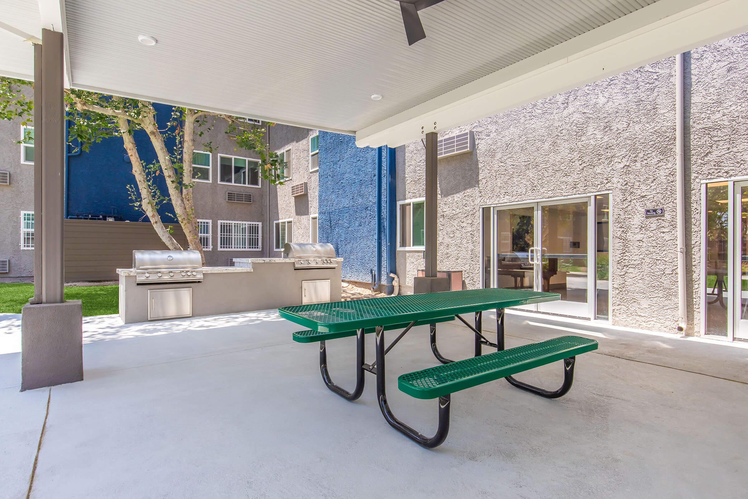 Outdoor communal area featuring a green picnic table under a covered patio. In the background, there is a barbecue grill setup and well-maintained grass. Surrounding buildings have textured gray exteriors with large windows, providing a welcoming atmosphere for gatherings.