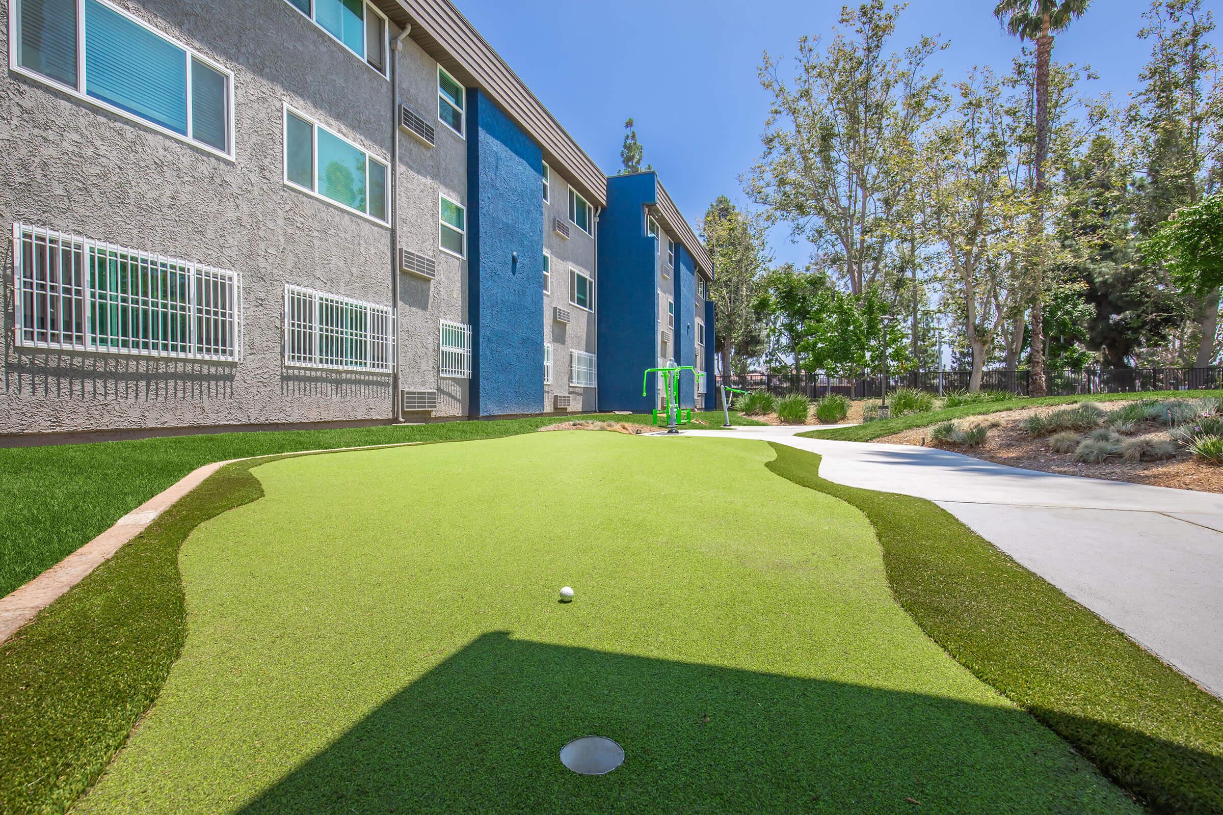 A landscaped outdoor area featuring a small putting green with a flag hole, surrounded by artificial turf. In the background, there are modern apartments with large windows and a few trees. A playground structure is visible nearby, adding to the recreational atmosphere.