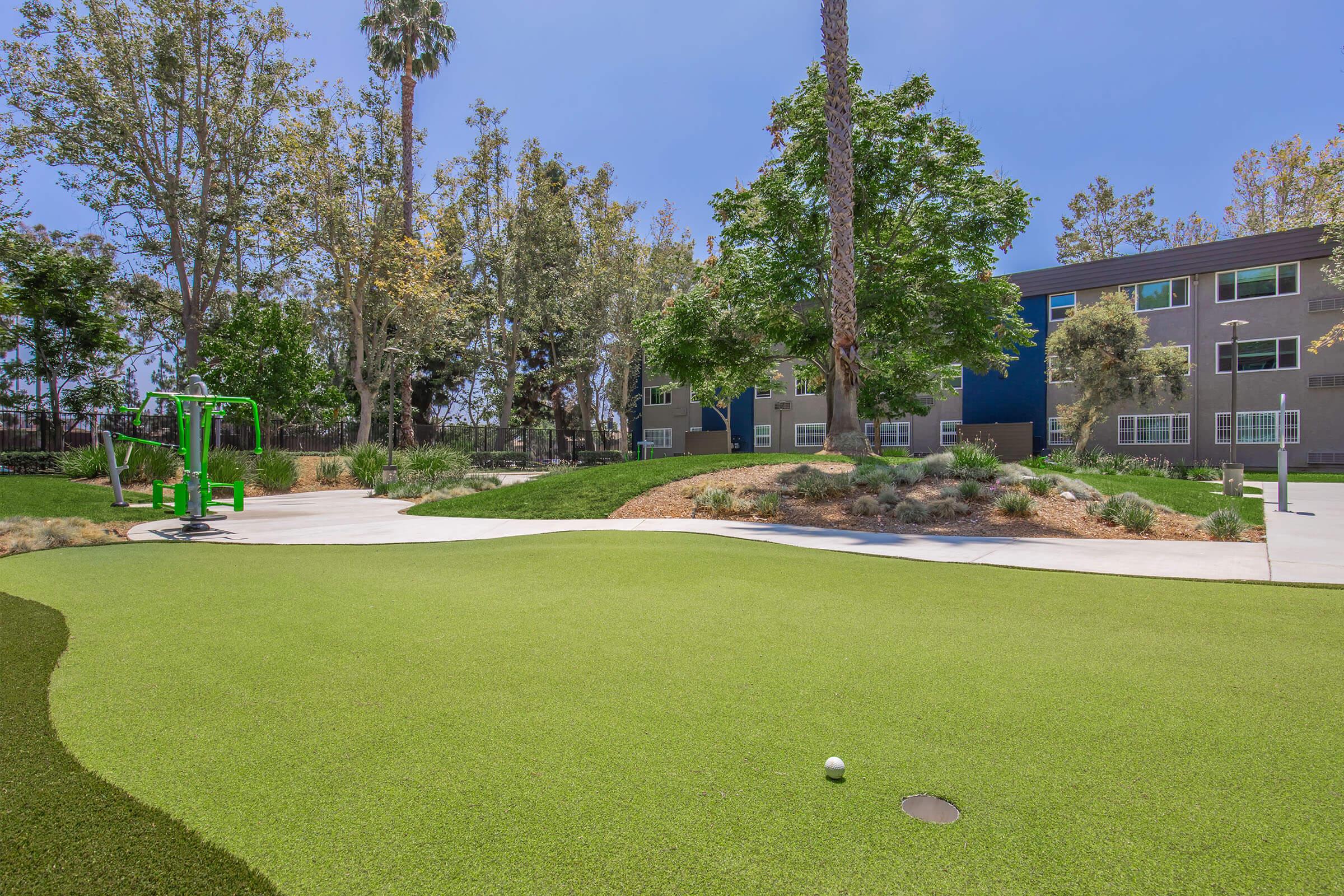 A well-maintained outdoor recreational area featuring green artificial grass, exercise equipment, and landscaped trees. In the background, there are modern residential buildings surrounded by greenery and pathways. The sky is clear, enhancing the inviting atmosphere of the space.