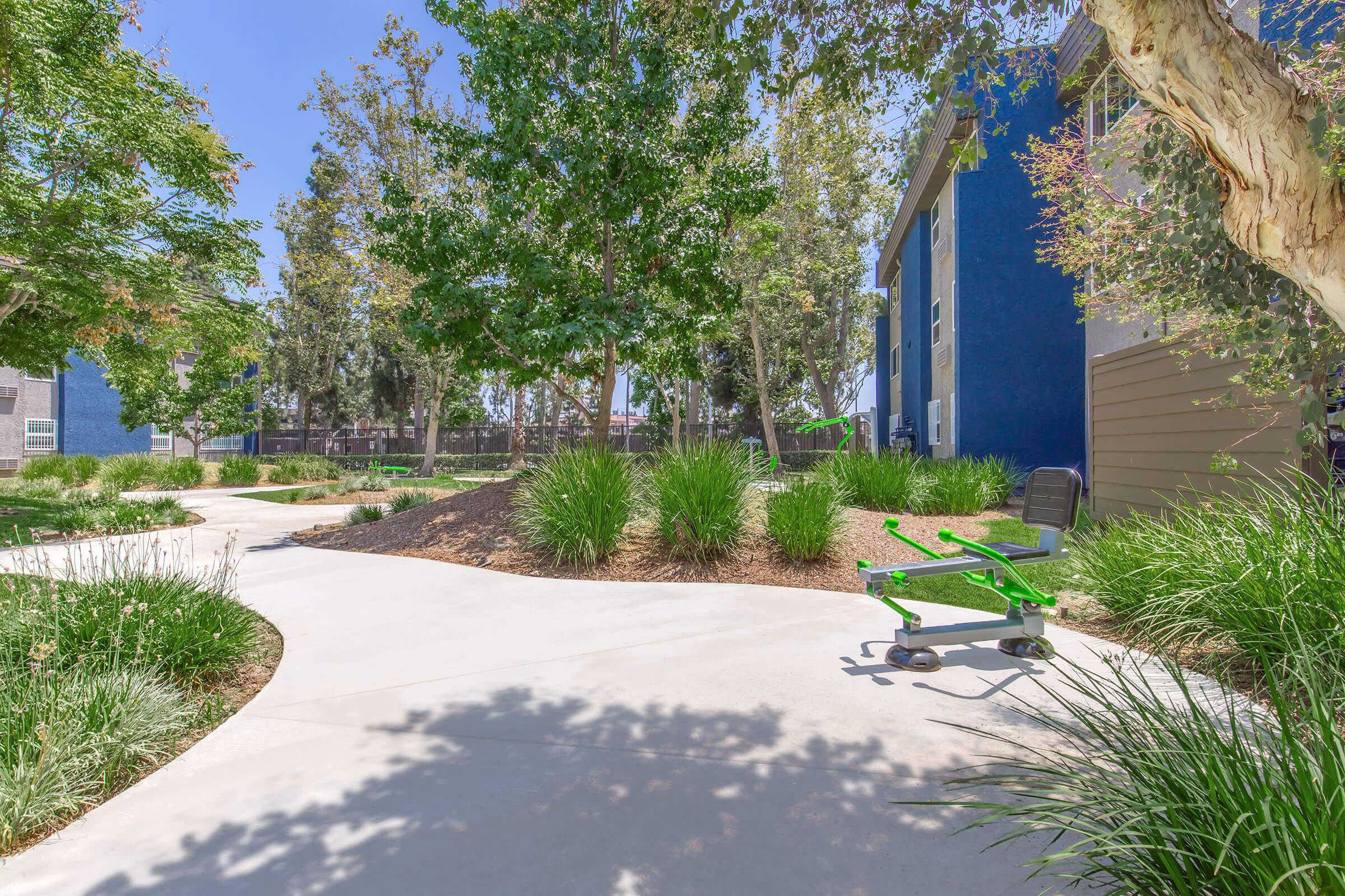 A landscaped pathway surrounded by greenery, leading through a park-like setting. In the foreground, there's a green exercise station with a bench, and two blue apartment buildings are visible in the background under a clear blue sky.