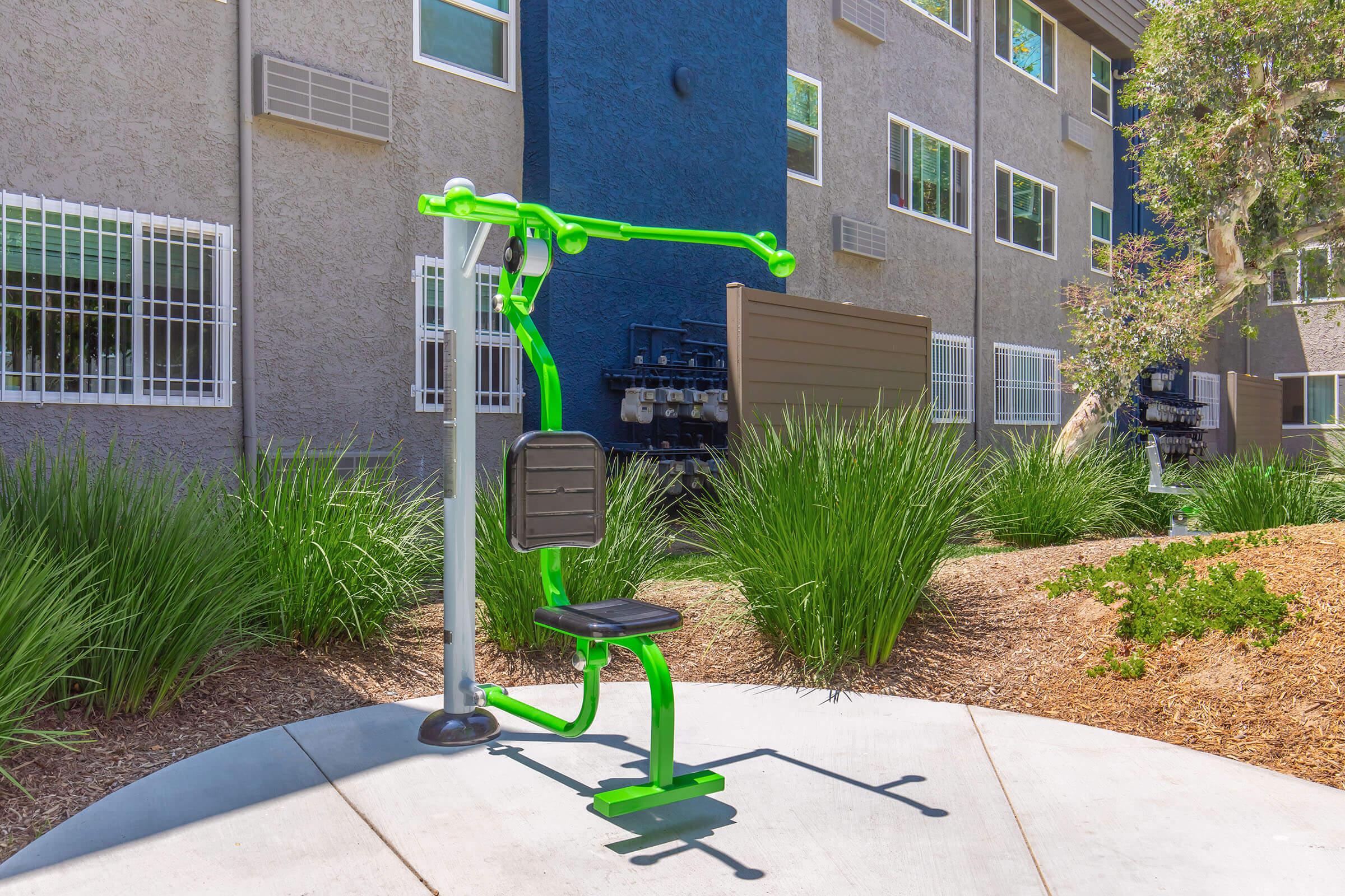Outdoor fitness equipment featuring a green exercise machine designed for upper body workouts, set on a concrete pad surrounded by decorative landscaping and residential buildings in the background. Ideal for promoting physical activity in communal spaces.