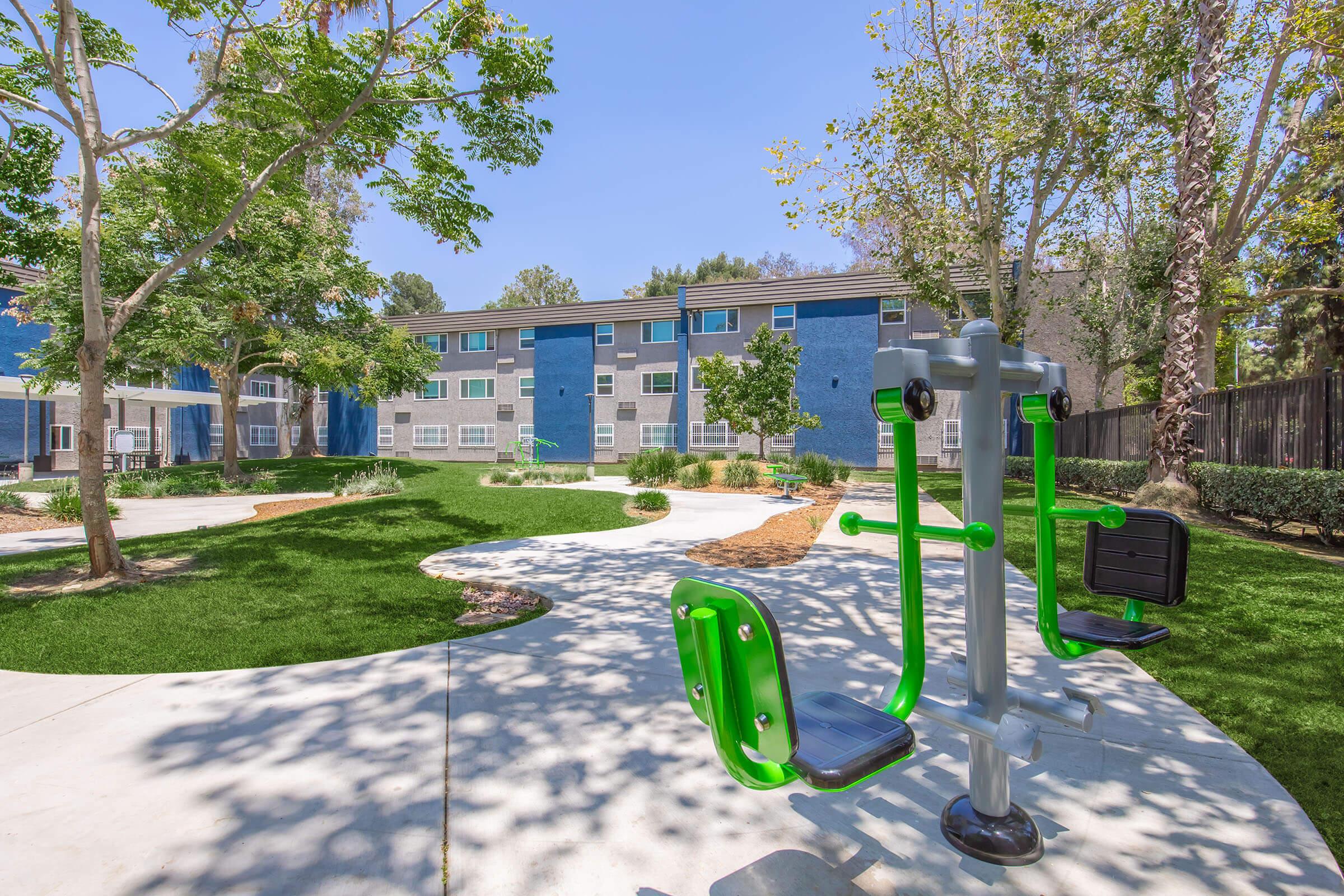 A vibrant outdoor exercise area featuring green and gray fitness equipment, surrounded by lush grass and trees. In the background, there are modern blue and gray buildings. The scene is bright and inviting, suggesting a community space for physical activity and recreation.