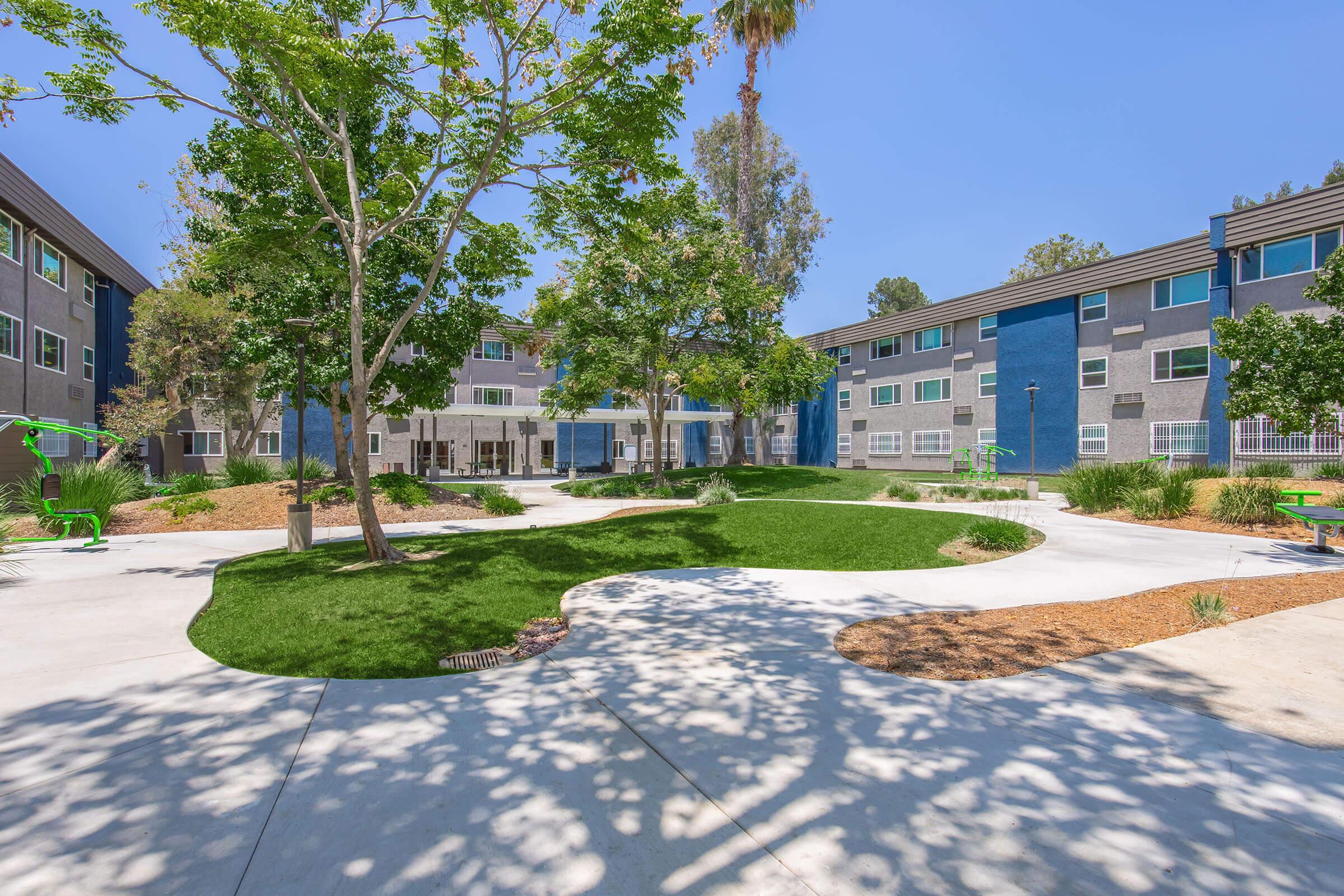 A landscaped courtyard featuring green grass, trees, and pathways. Surrounding buildings are painted in shades of gray and blue, with large windows. The scene is bright and sunny, creating a welcoming outdoor space for residents.