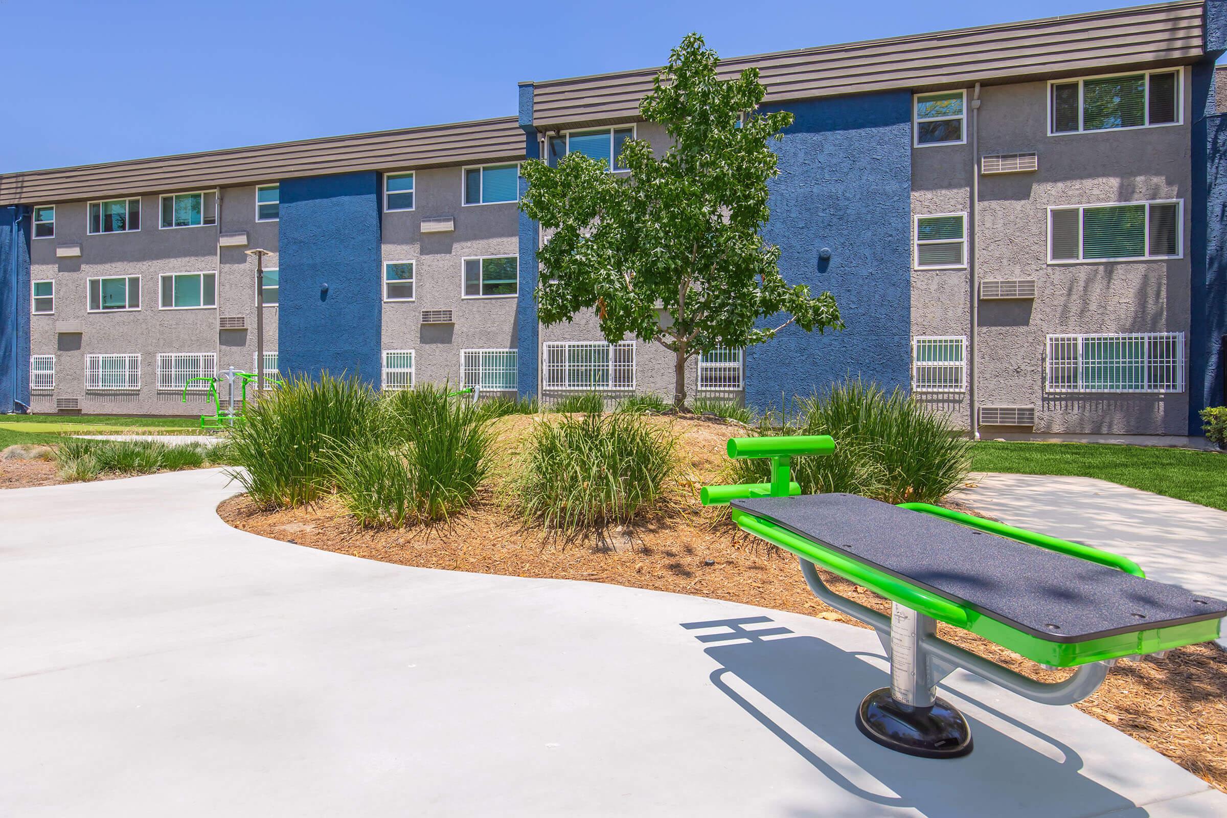 Outdoor fitness area with green exercise equipment, surrounded by neatly trimmed grass and shrubs. In the background, multicolored apartment buildings with large windows. The layout is designed for recreational activities in a sunny environment.