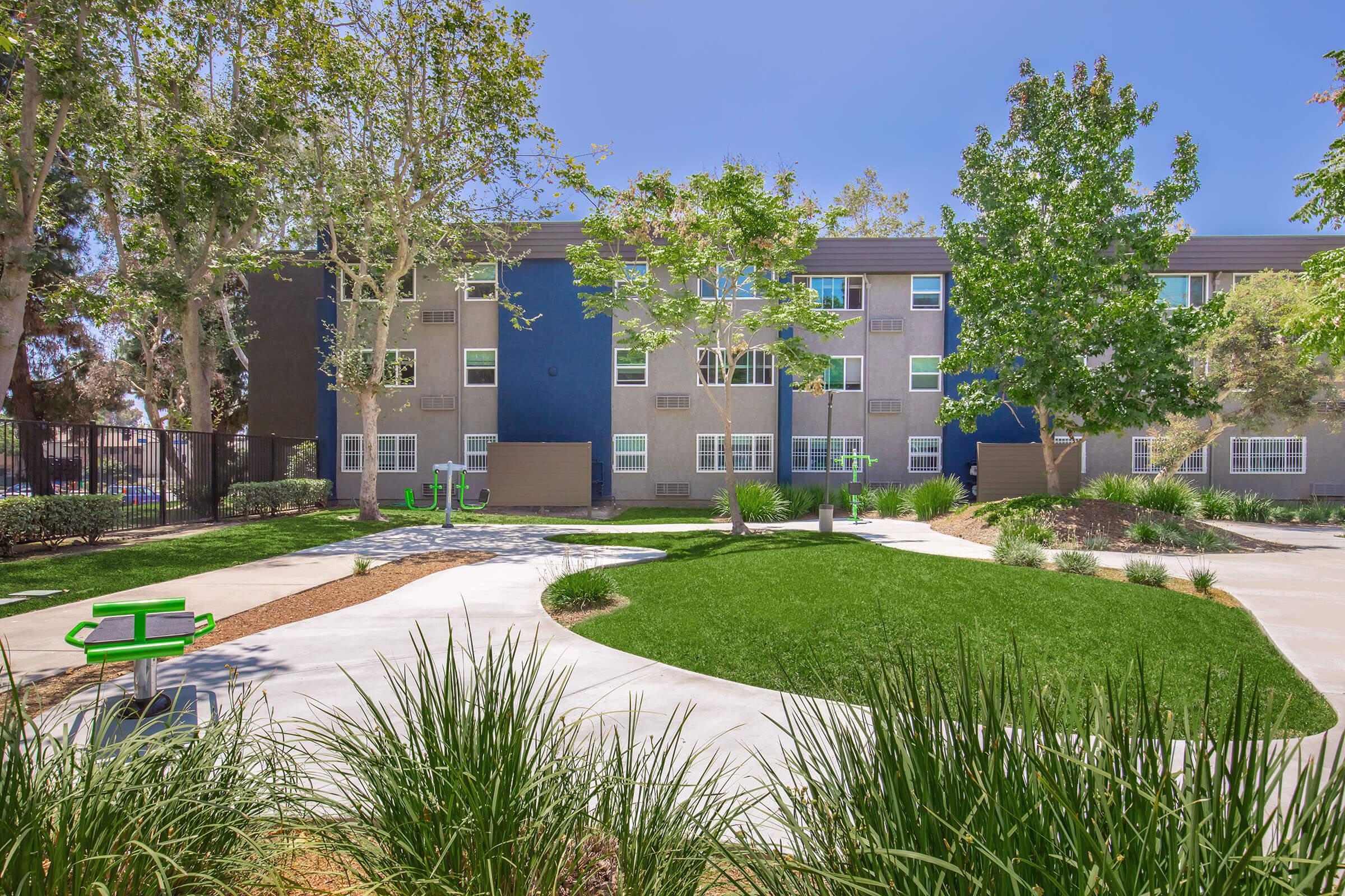 A landscaped outdoor area featuring a green lawn, walking paths, and trees. In the background, there is a modern multi-story building with a blue and gray facade. Two green benches are visible, providing a place to sit and enjoy the surroundings. The sky is clear and sunny.
