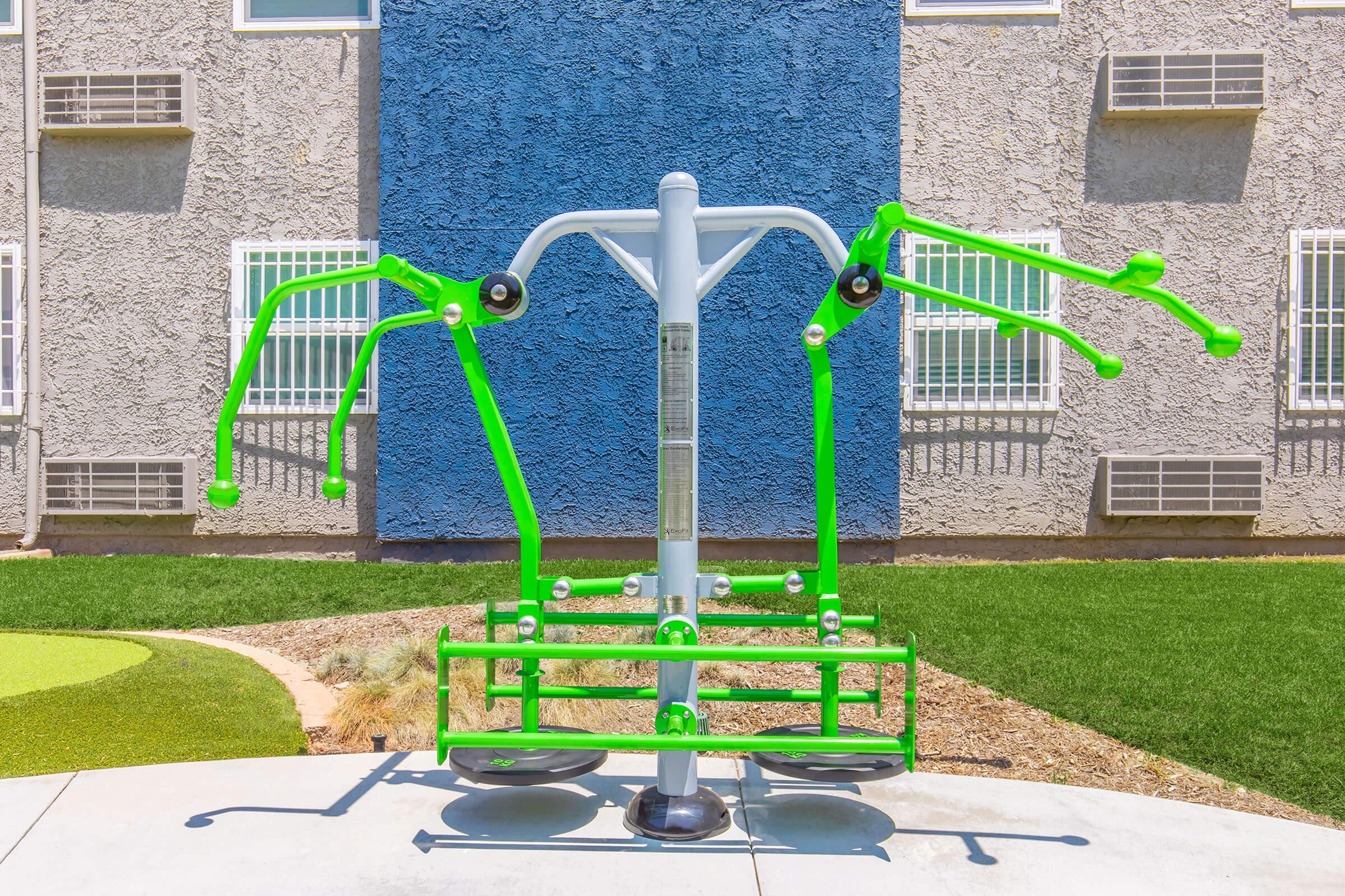 Outdoor fitness equipment with two green handles for arm exercises, positioned on a concrete base in a lawn area. Behind it is a textured gray wall with a blue accent. The setting is bright and sunny, ideal for outdoor workouts.