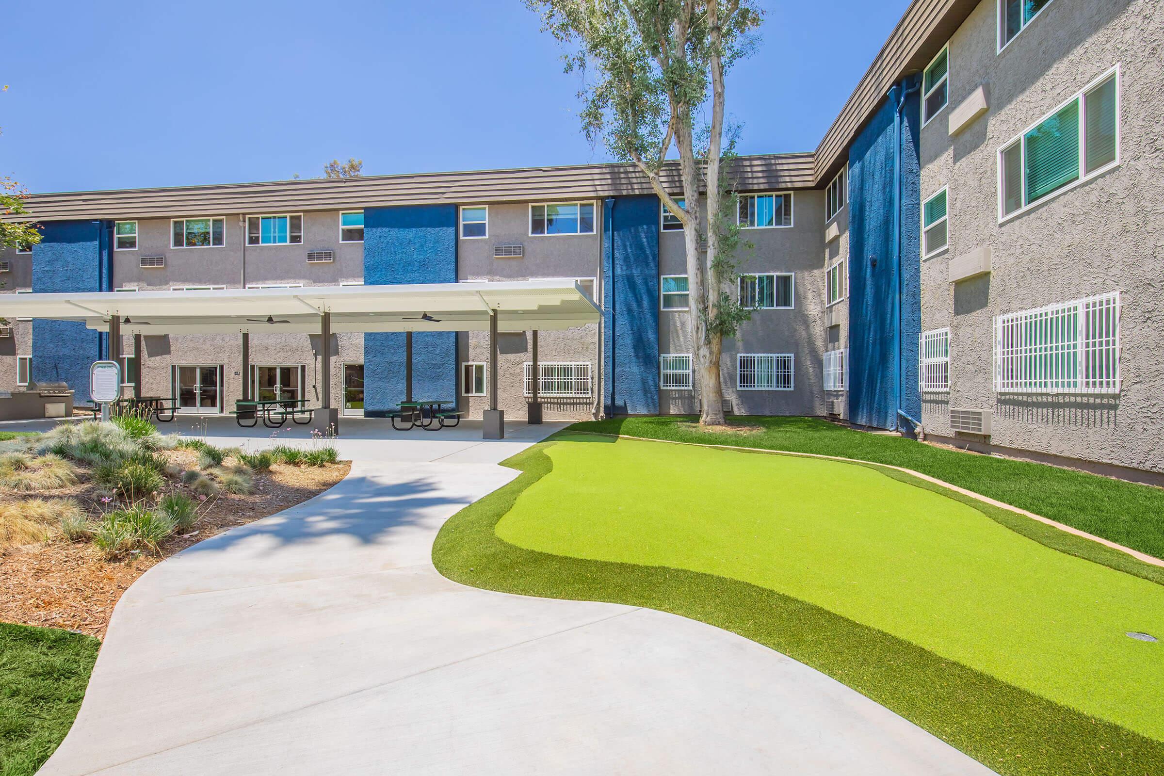 A modern residential building featuring a well-maintained lawn with artificial turf. The exterior is a mix of gray and blue colors, and there's a shaded outdoor seating area with tables. The sky is clear and blue, enhancing the inviting atmosphere of the space.