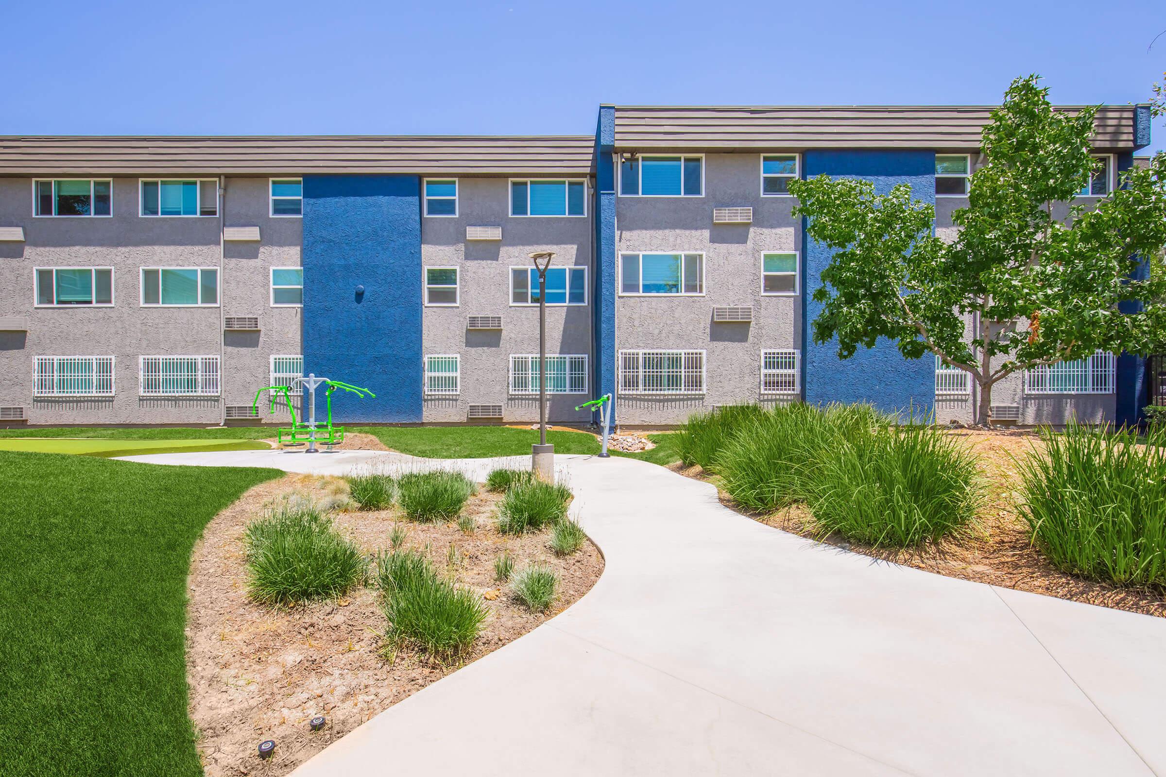 A modern building with a blue and gray exterior, featuring multiple windows. In the foreground, there is a landscaped area with green grass, a winding concrete pathway, and small shrubs. Outdoor fitness equipment is visible in the garden area, under a clear blue sky.