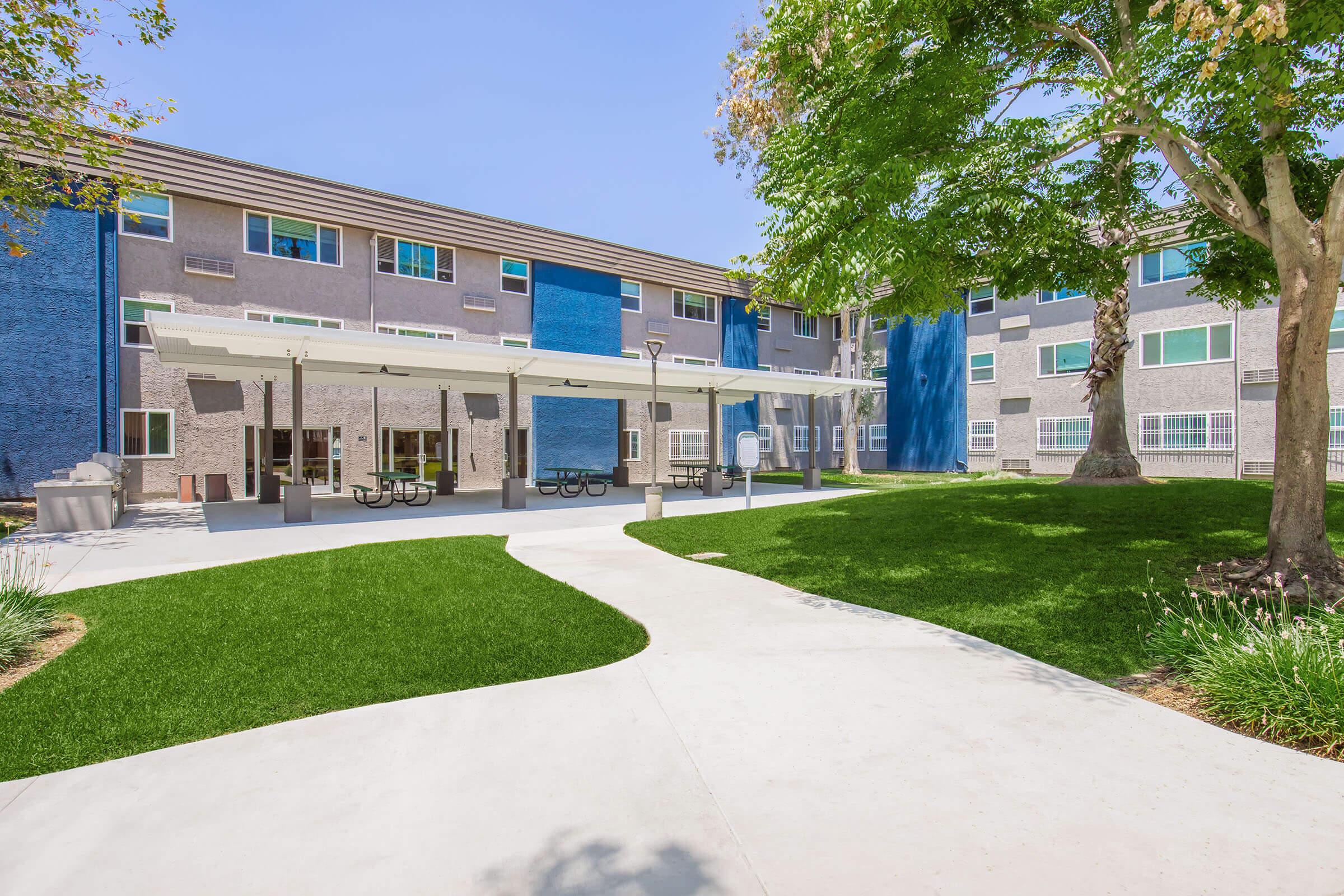A sunny courtyard featuring green grass and a concrete pathway. There are shaded picnic areas with tables under a large canopy, surrounded by modern multi-story buildings with blue and neutral-colored exteriors. Trees provide additional shade in the outdoor space.