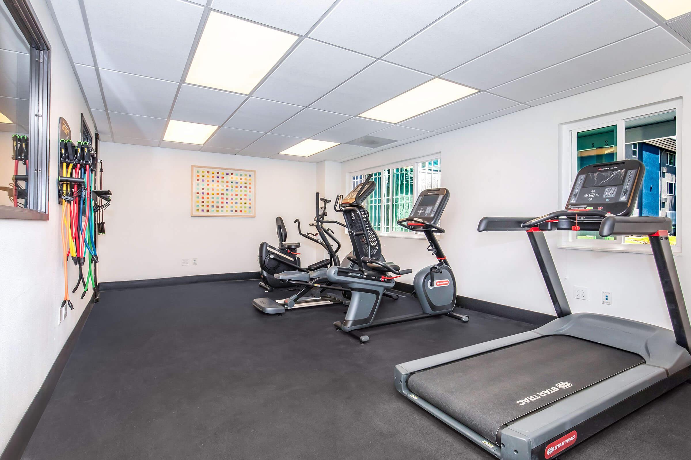 A bright and modern fitness room featuring various exercise equipment, including treadmills and stationary bikes. There are fitness bands mounted on the wall and large windows providing natural light, creating a motivating workout environment. The floor is covered with black rubber flooring for safety.