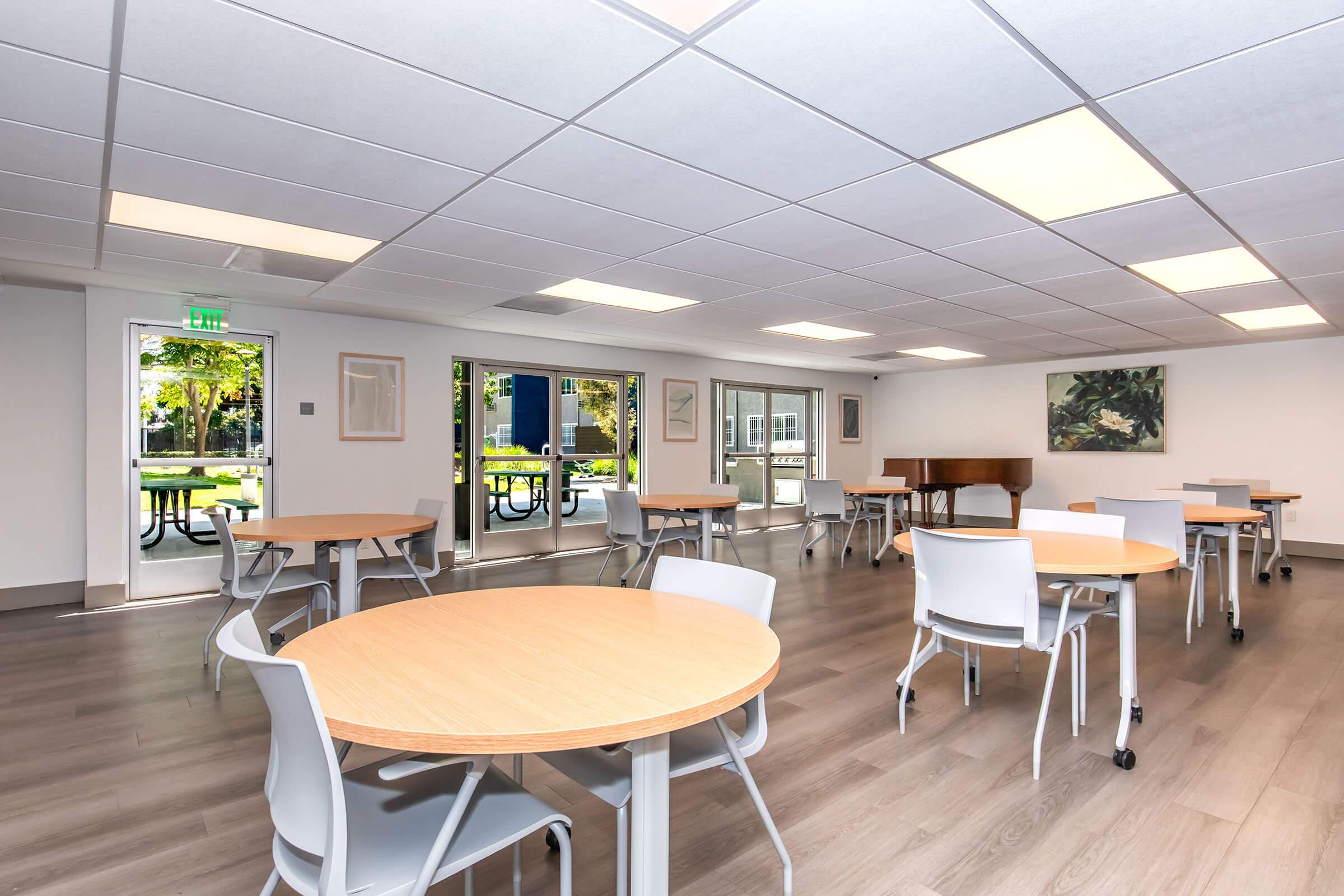A spacious, well-lit room featuring several round wooden tables and white chairs. Large windows allow natural light to fill the space, with views of greenery outside. A piano is positioned against the wall, and artwork is displayed on the walls, creating a welcoming environment suitable for gatherings or relaxation.
