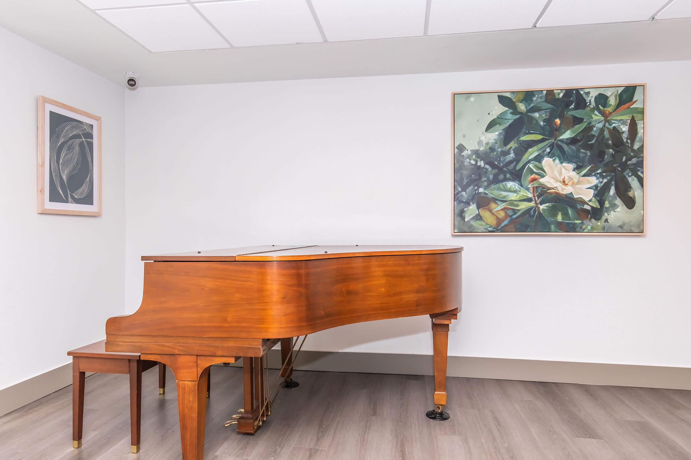 A polished wooden grand piano is positioned in a well-lit room with modern decor. It sits on a light hardwood floor, accompanied by a framed artwork featuring a floral theme on the wall and another abstract piece nearby. A ceiling camera is visible in the corner.
