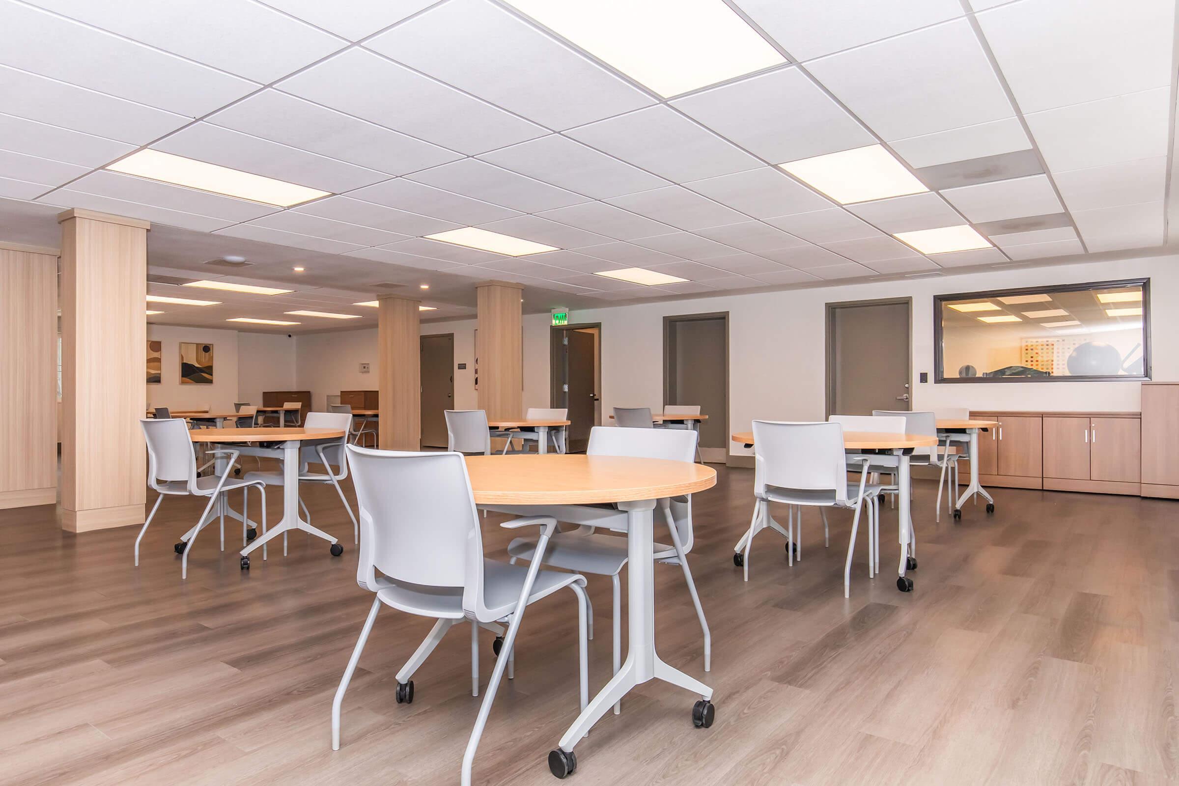 A spacious, modern classroom or meeting room featuring multiple round tables with light-colored wooden tops and gray chairs. The floors are light brown, and the ceiling has fluorescent lighting. There are windows allowing natural light, and the walls are painted in neutral tones.