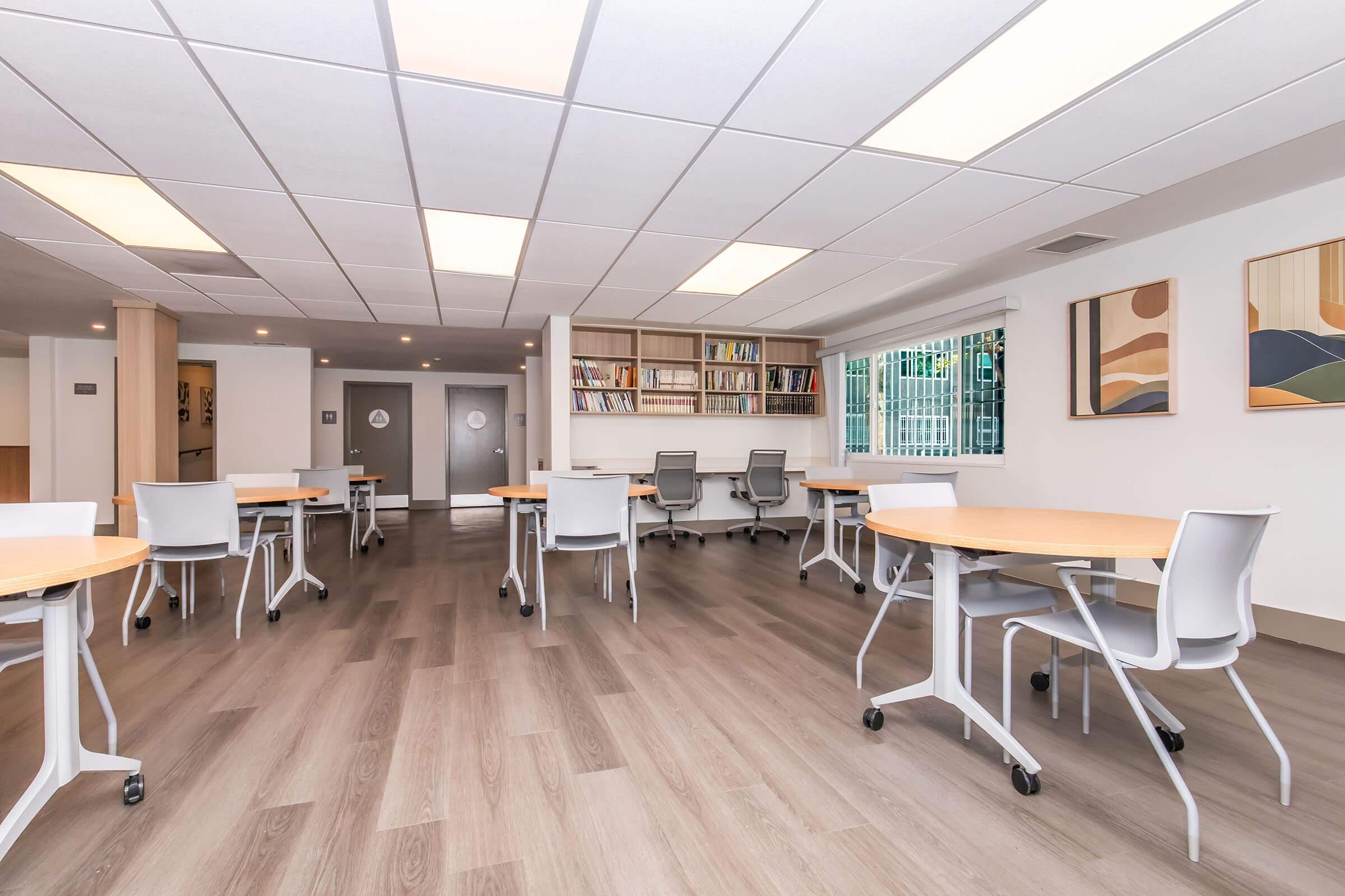 A spacious, modern study area featuring several round wooden tables with white chairs. Bookshelves line one wall, filled with books and decorative items. Large windows allow natural light to flood the space, and a calming color palette enhances the inviting atmosphere.
