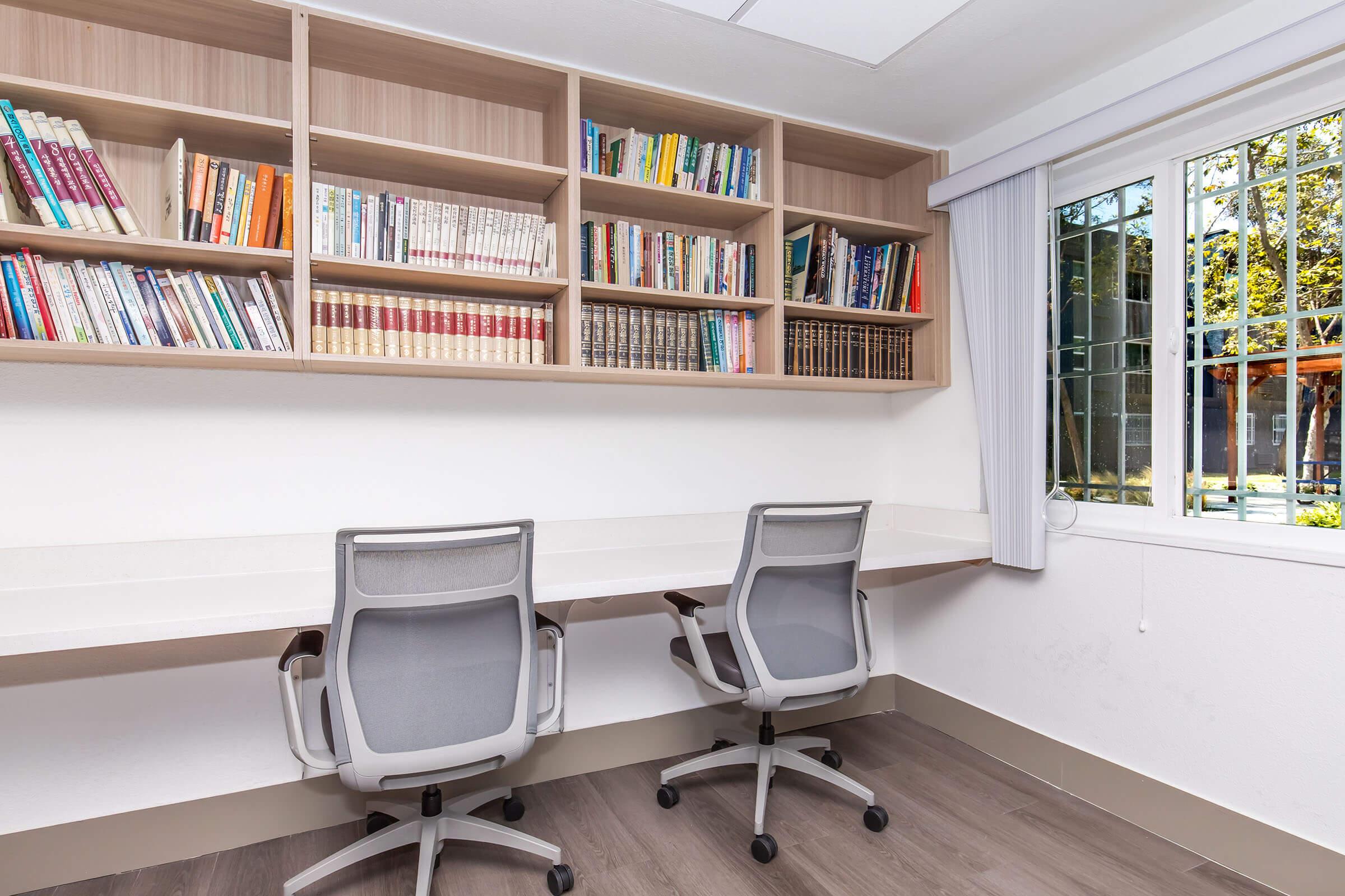 A tidy workspace featuring two chairs and a long desk. Above the desk, there is a wooden bookshelf filled with various books. A window provides natural light, revealing a glimpse of an outdoor area with trees and a patio. The overall atmosphere is clean and organized, suitable for studying or working.