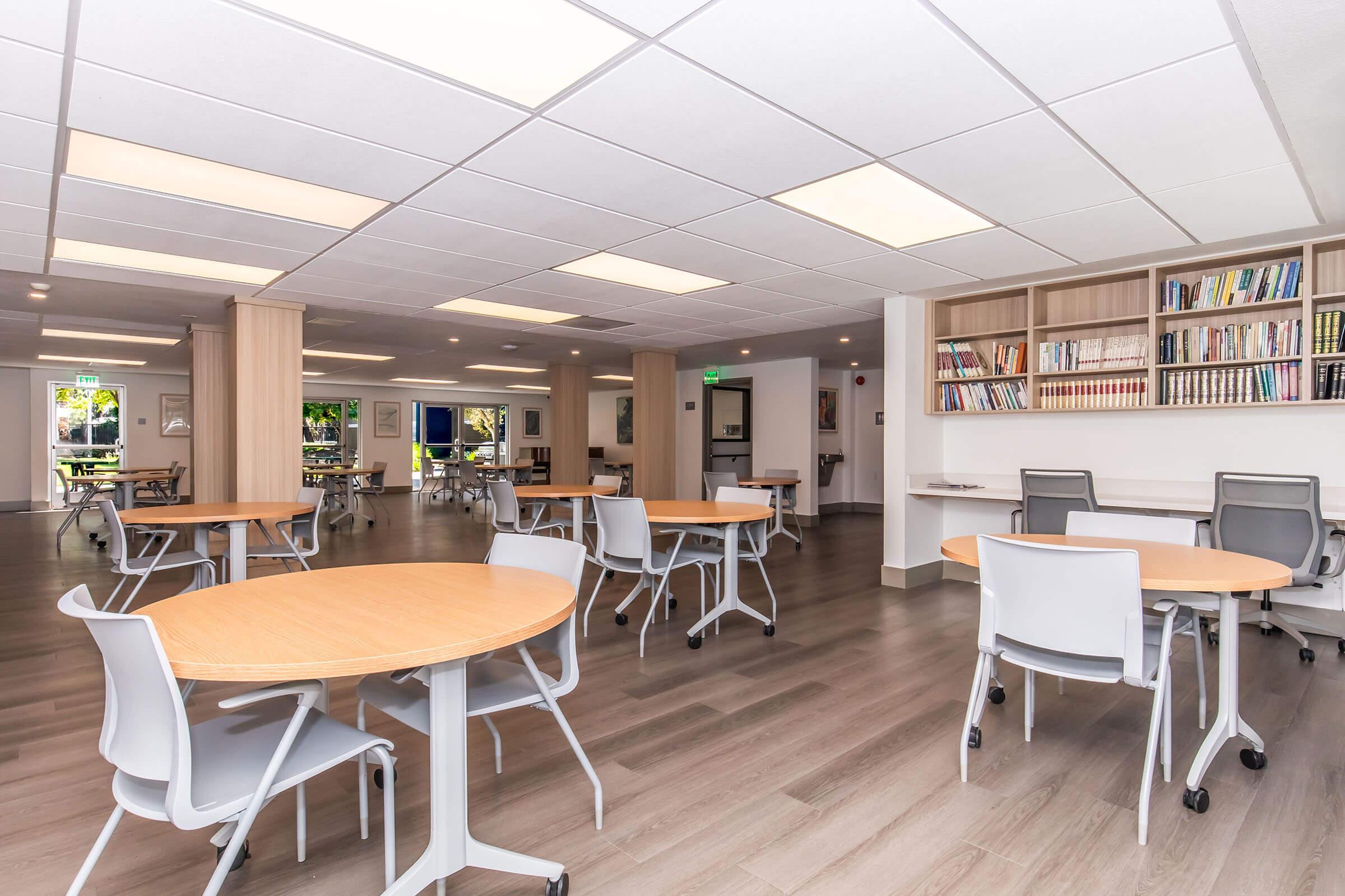 Interior of a modern, spacious communal area featuring several round wooden tables with gray chairs. The room is well-lit with overhead lighting and has large windows allowing natural light. Bookshelves filled with books are visible along one wall, contributing to a warm and inviting atmosphere.