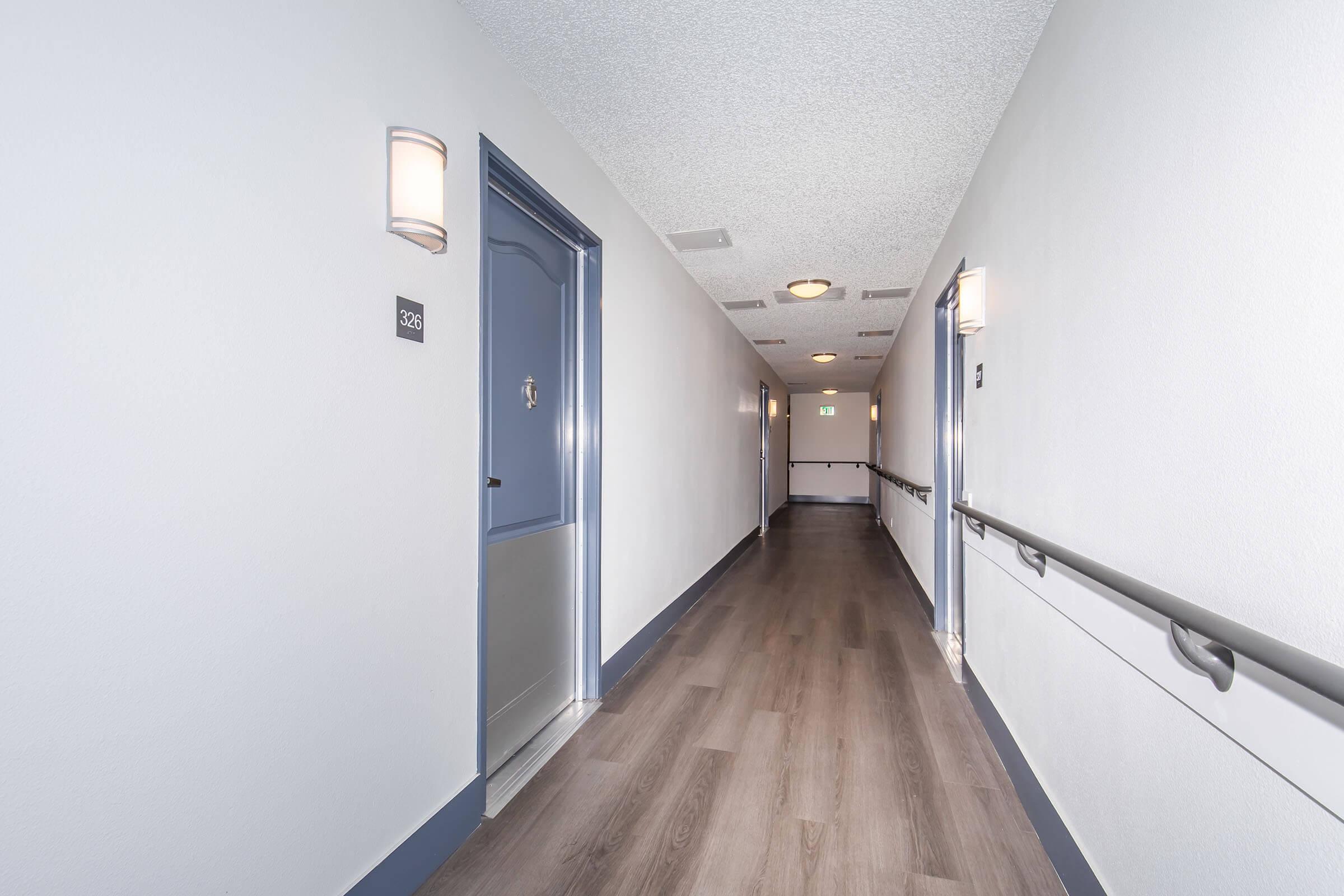 A clean, well-lit hotel corridor with slight gray walls and wooden flooring. On the left, there is a blue door labeled "326" and on the right, there are other doors and wall-mounted lights. The hallway appears modern and inviting, with a smooth railing along one side.