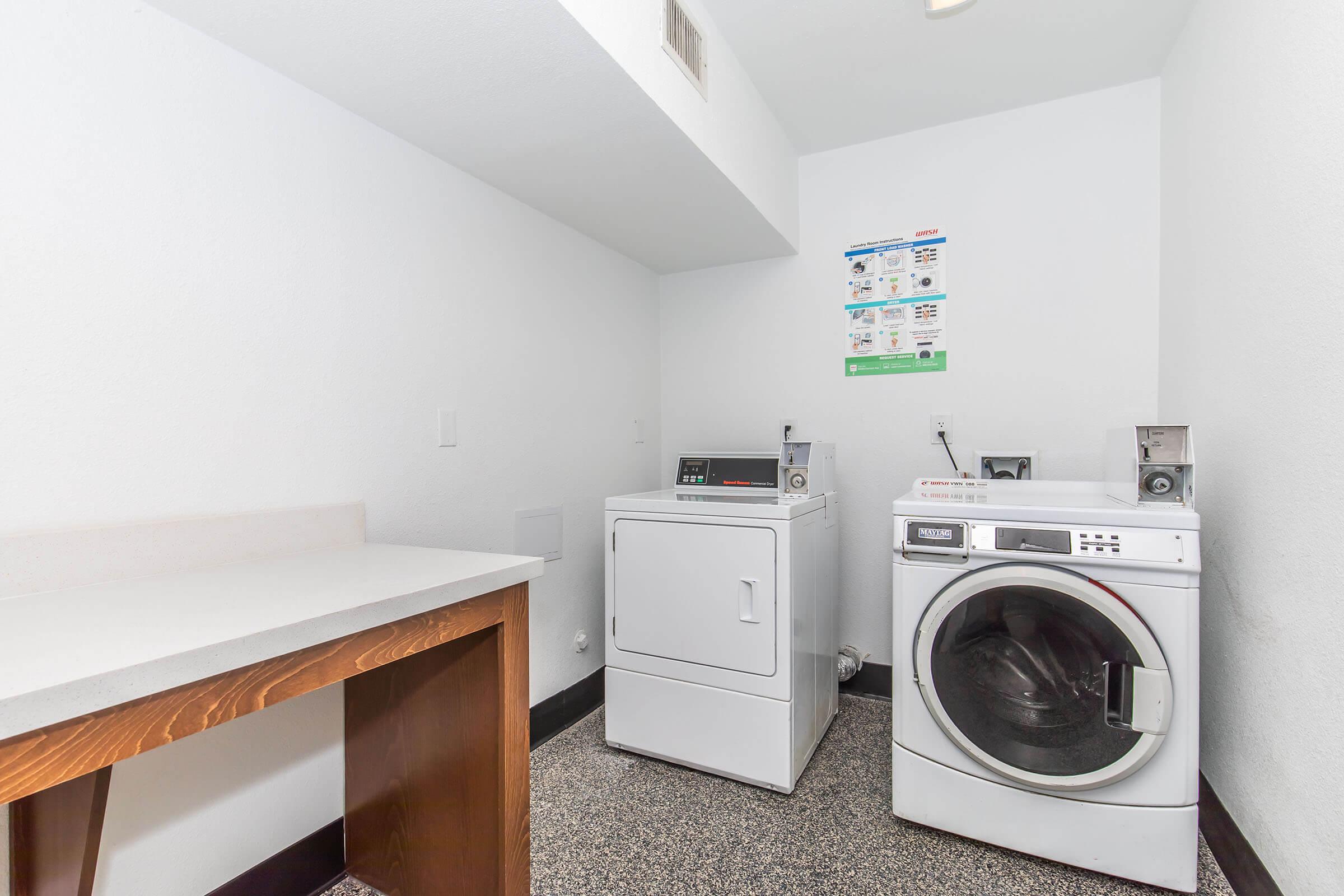 A laundry room featuring a white washing machine and a dryer. There is a countertop for folding clothes and a wall with informational posters. The floor is dark and the walls are white, creating a clean and organized appearance.
