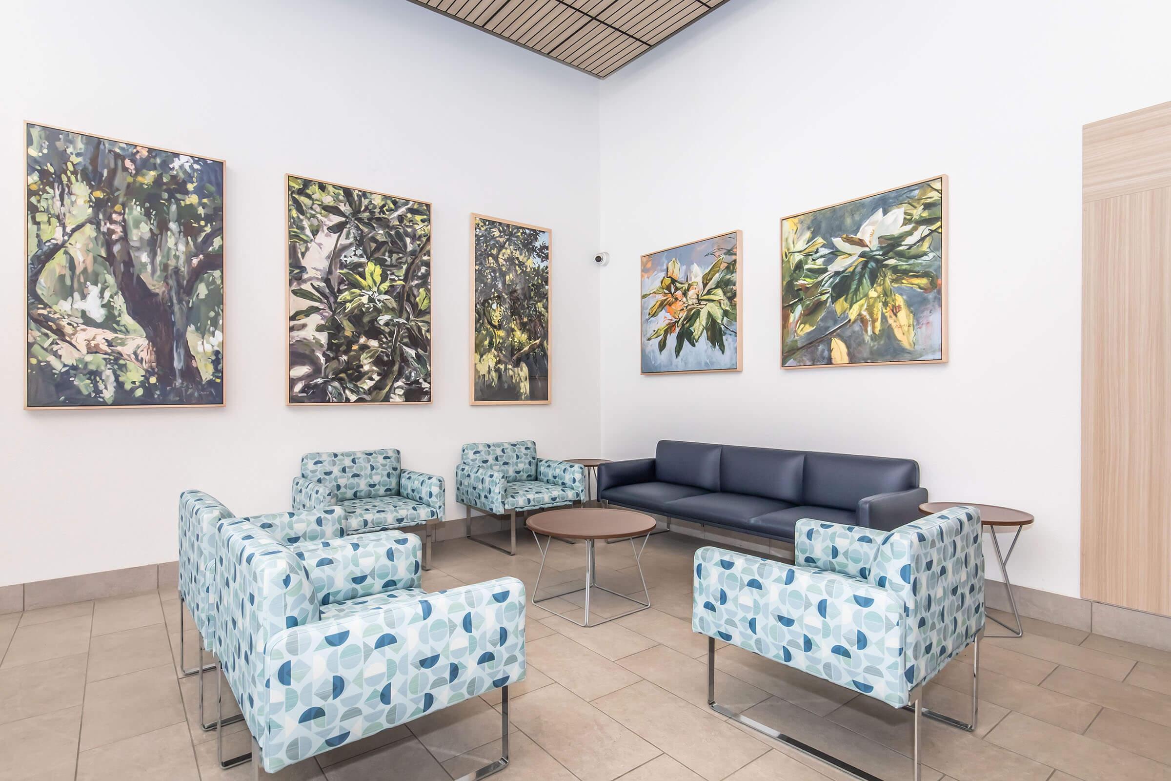 A modern waiting area featuring light-colored tiled flooring, a dark blue sofa, and four patterned chairs with circular designs. There is a round table in the center, and the walls are adorned with five large framed artworks depicting abstract nature scenes. The space is well-lit and inviting.