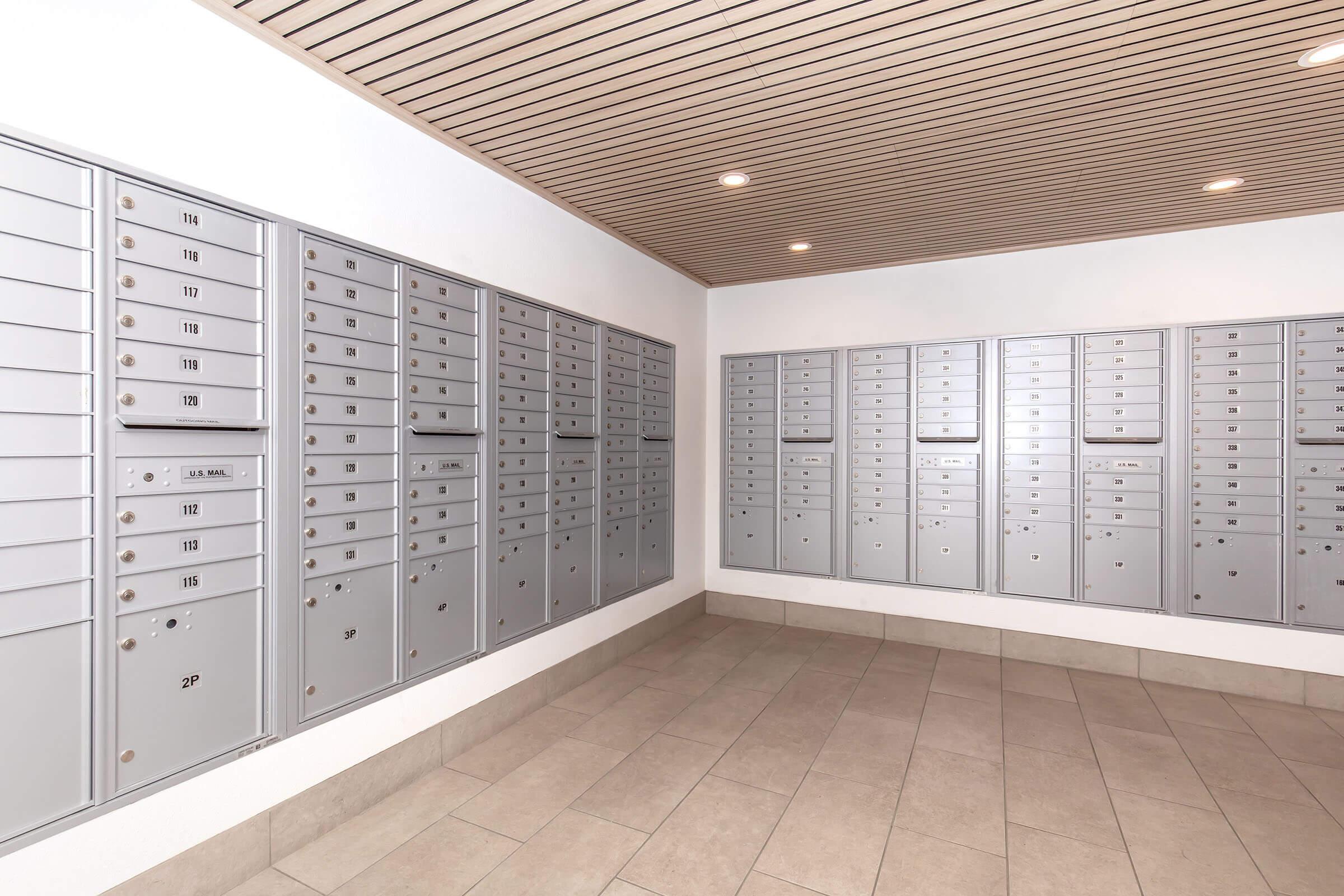 A modern mailroom with multiple metal mailboxes mounted on the wall, arranged in two sections. The interior features neutral-colored walls and flooring, with a wooden ceiling. Bright lighting illuminates the space, creating a clean and organized environment for postal services.