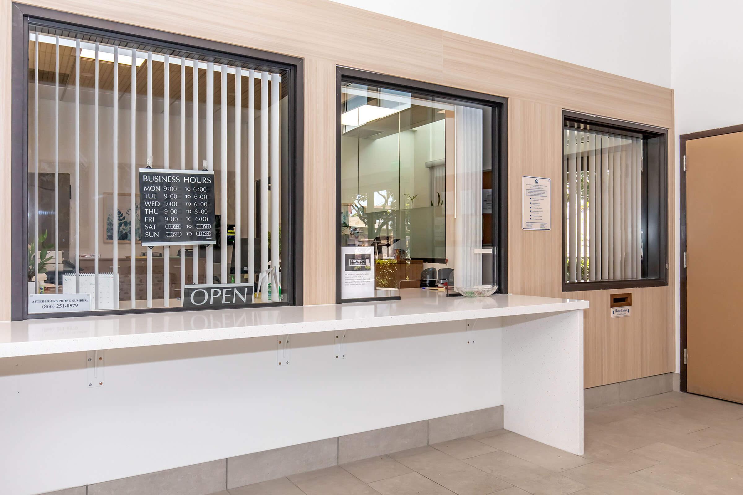 Reception area with a service counter and large windows featuring vertical blinds. A sign indicating business hours is displayed, with an "OPEN" sign visible. The room appears well-lit and modern, with a neutral color palette and a door on the right side.