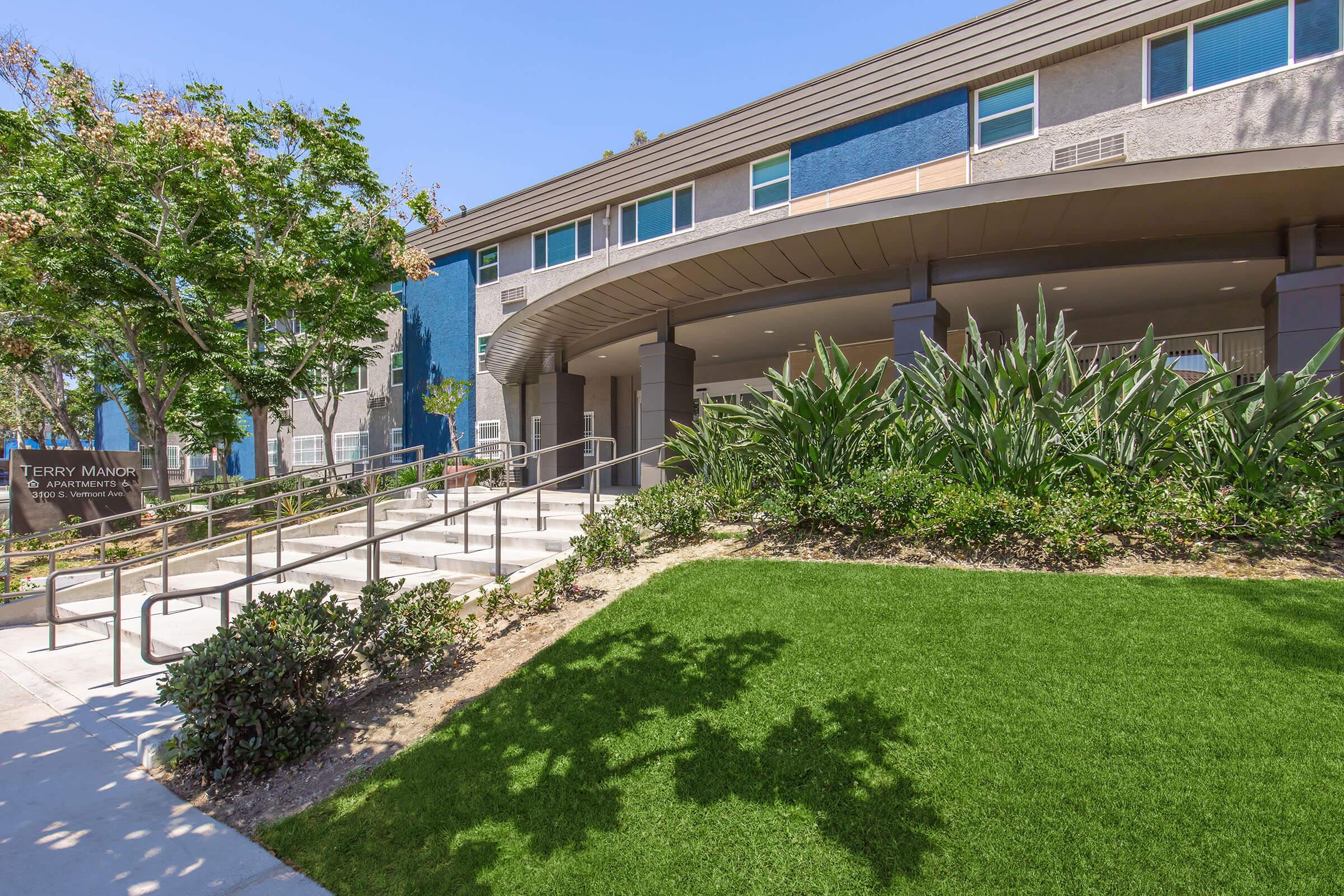 A modern building with large windows and a welcoming entrance, featuring a curved walkway with railings. Surrounding the building are well-maintained green lawns and tropical plants, creating a pleasant outdoor space in a sunny environment. The design is accessible and inviting.