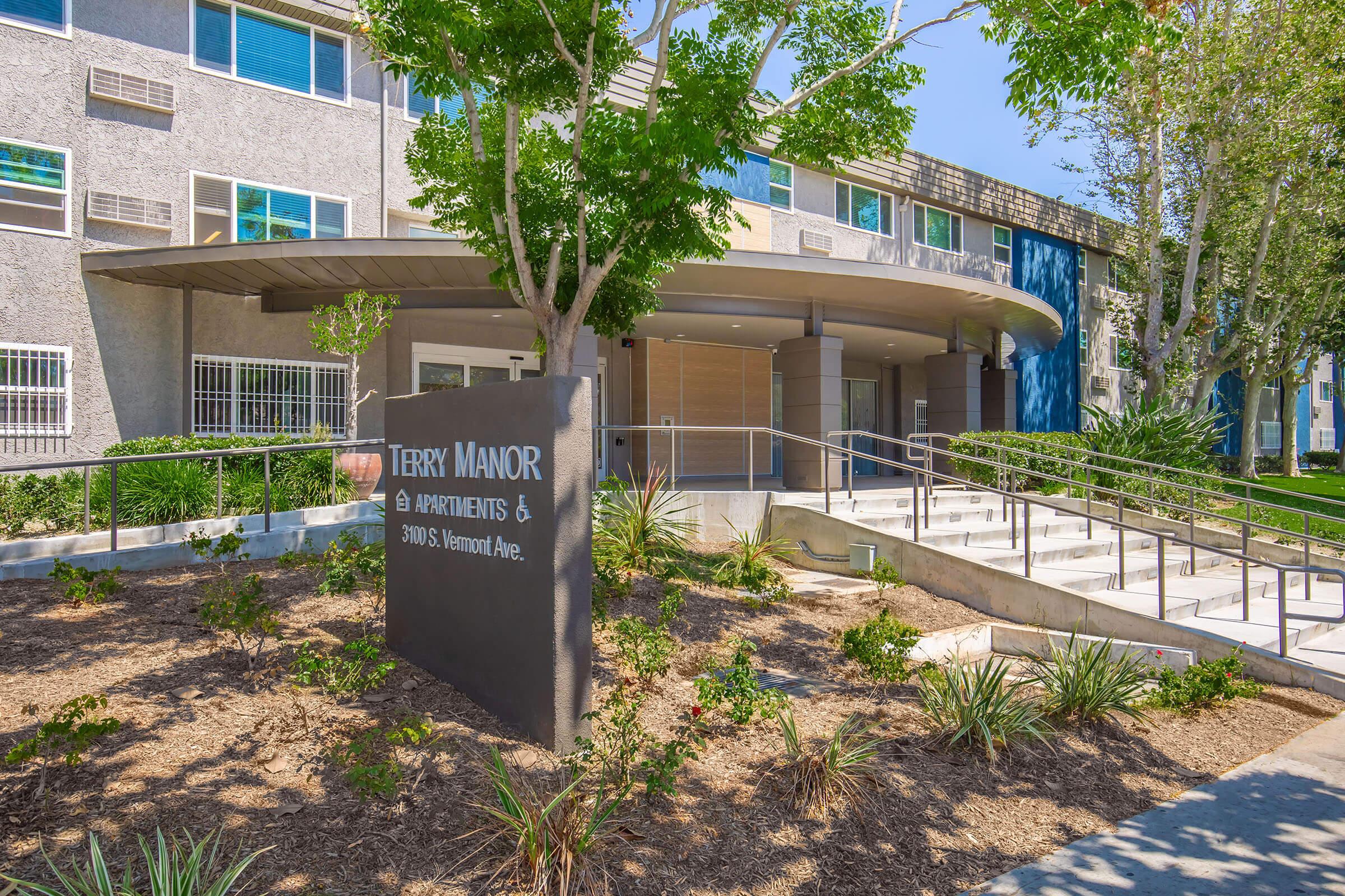 Exterior view of Terry Manor Apartments featuring a modern entrance with steps and a landscaped area. The sign displaying "Terry Manor Apartments" is prominently displayed in front. Surrounding greenery adds a welcoming touch to the building's facade.