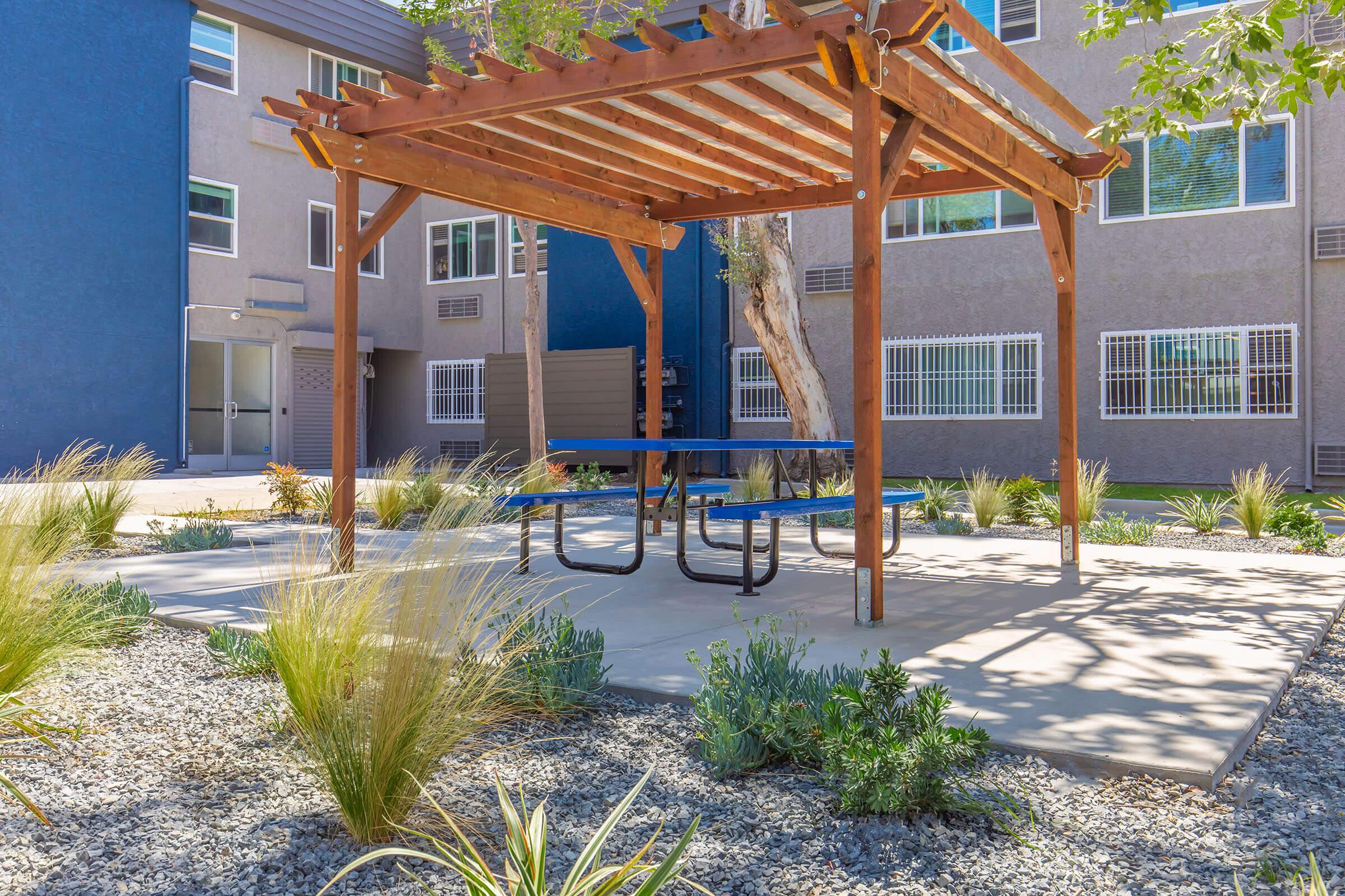 A wooden pergola with a blue picnic table underneath, surrounded by decorative gravel and low-maintenance plants. In the background, there are modern apartment buildings with large windows, creating a serene outdoor space for relaxation.