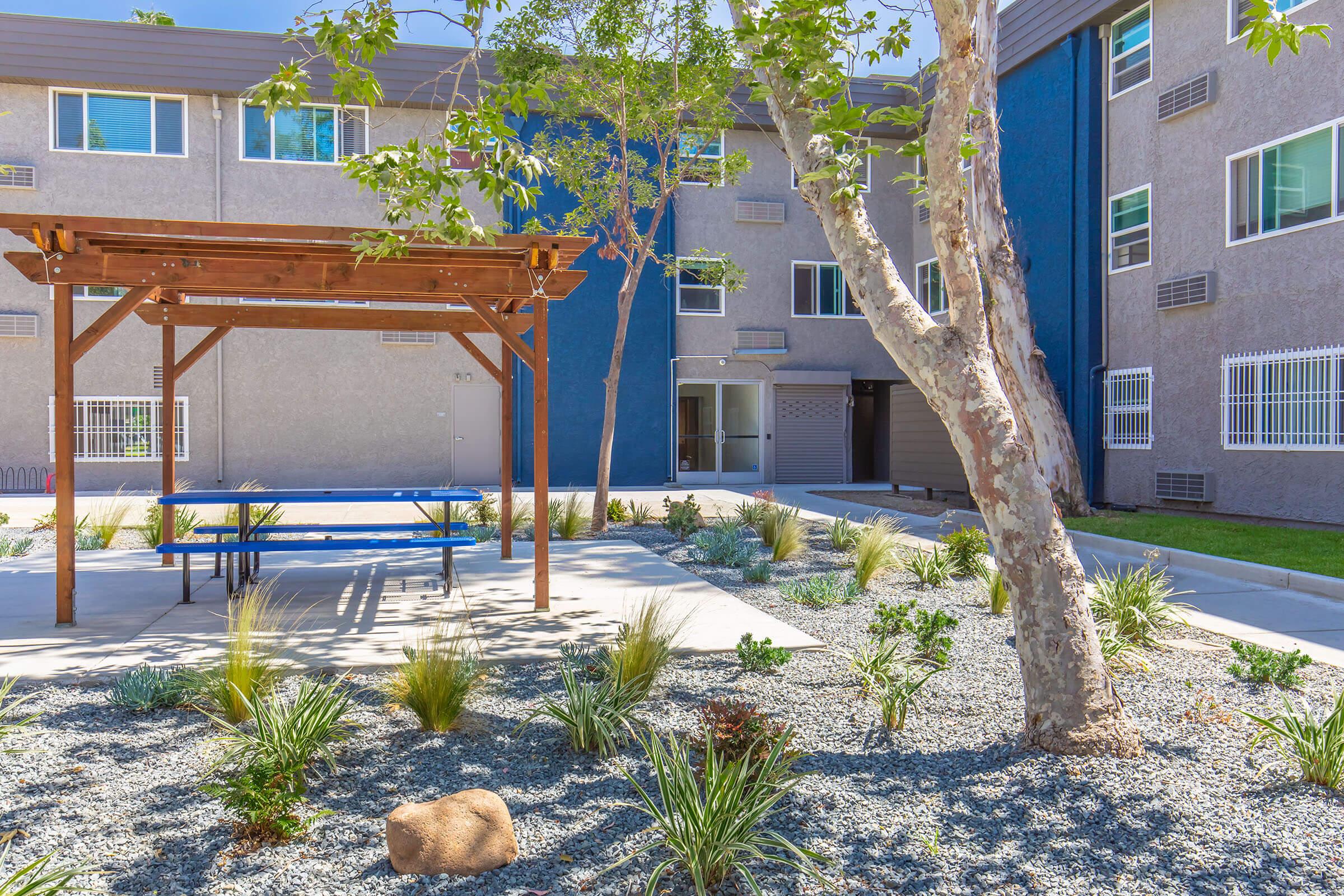 A landscaped courtyard featuring a wooden pergola with a blue picnic table underneath, surrounded by gravel and greenery. In the background, there are modern apartment buildings with large windows and a blue accent wall. Sunlight brightens the scene, creating a welcoming outdoor space.