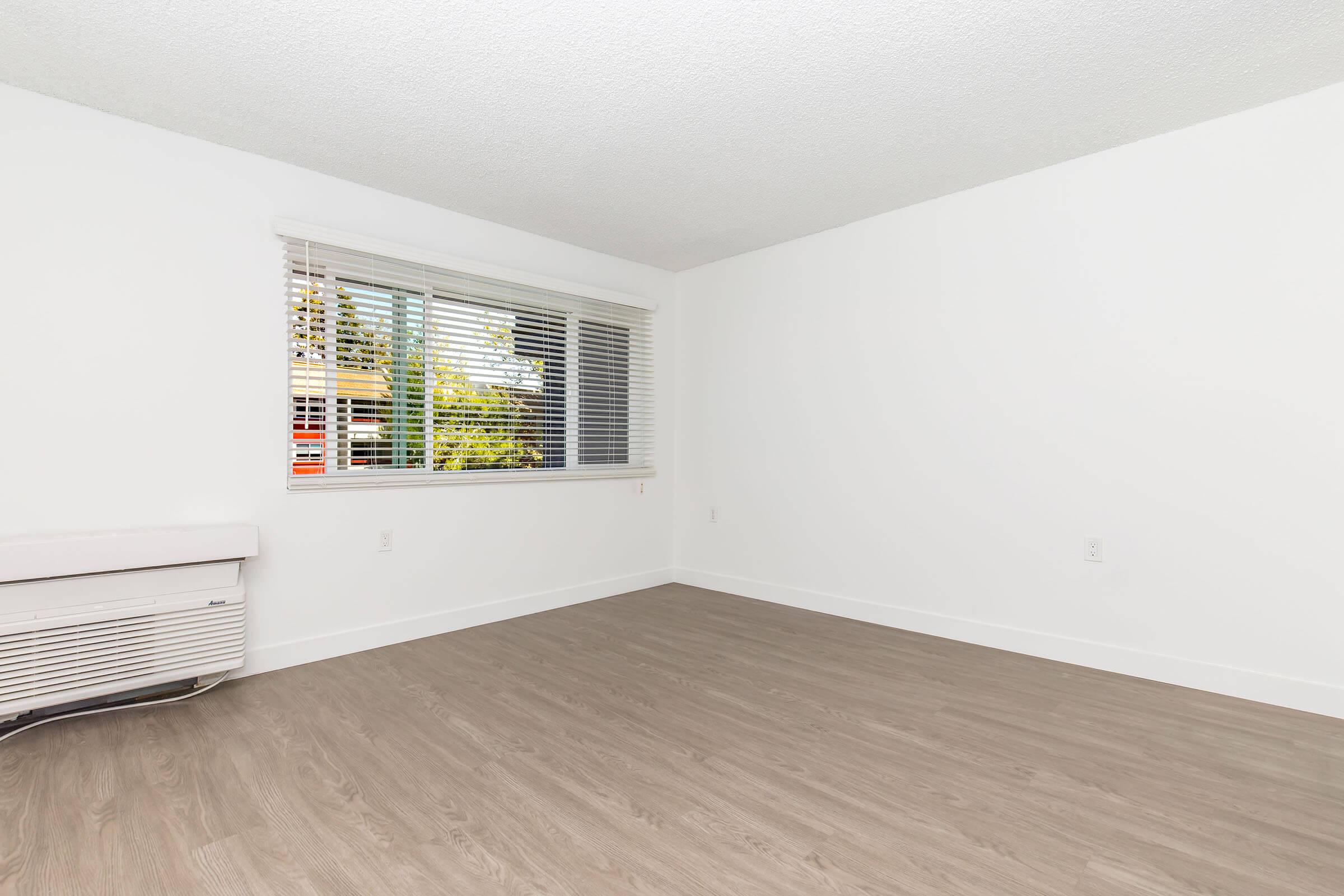 A clean and empty room with light-colored walls and a light wood laminate floor. It features a large window with blinds, allowing natural light to enter. An air conditioning unit is visible on the left side of the wall. The overall atmosphere is bright and minimalistic.