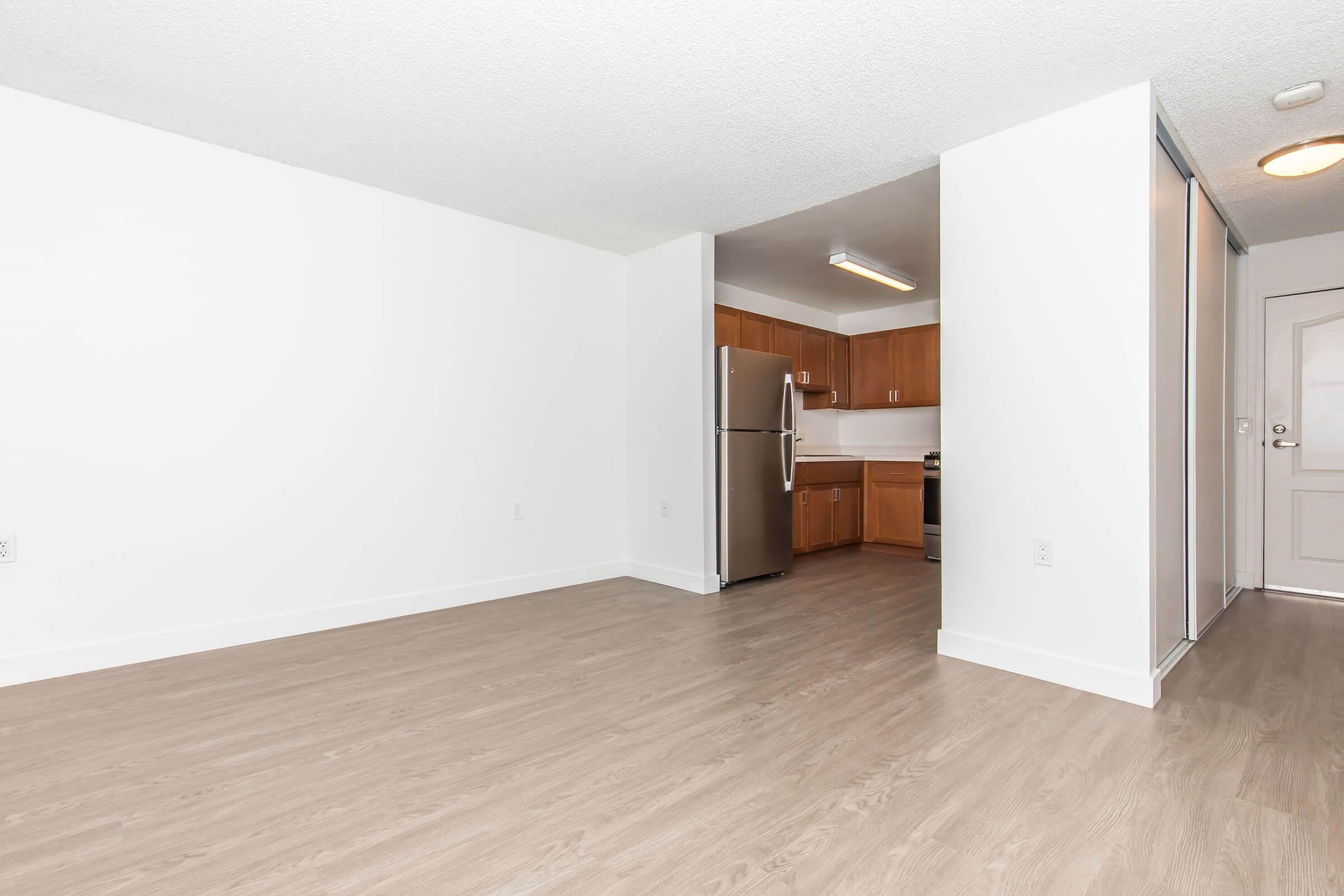 A modern, empty apartment interior featuring light-colored walls and hardwood flooring. The kitchen area is visible in the background with wooden cabinets, a stainless steel refrigerator, and stainless steel appliances. The space is bright and well-lit, with a door leading to the entrance on the right.