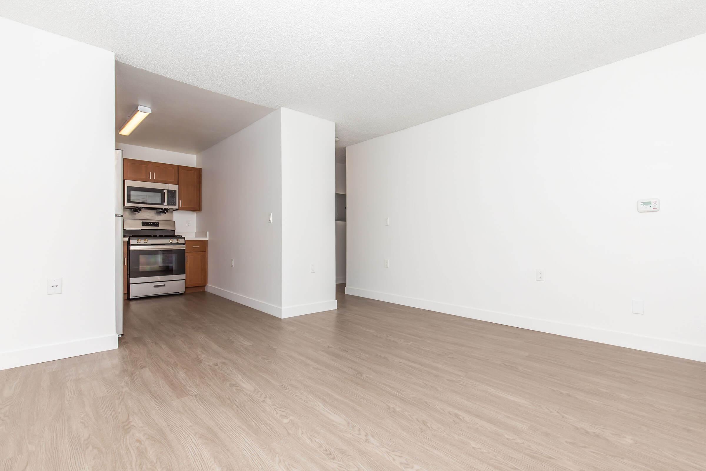 A spacious, empty living area featuring light-colored walls and wooden flooring. To the left, a modern kitchen with stainless steel appliances including a stove, microwave, and refrigerator. The open layout offers ample natural light and a clean, minimalist design.