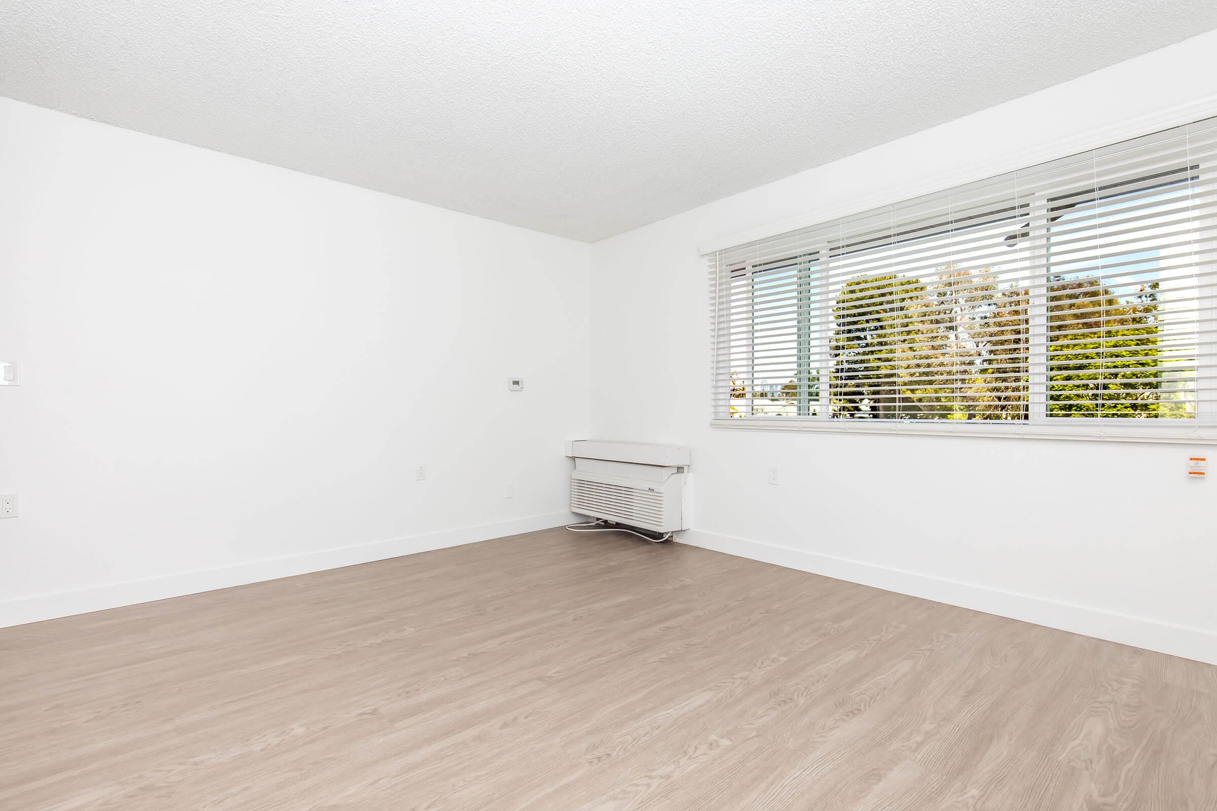 A bright, empty room with white walls and a large window covered by blinds. There's a white air conditioning unit on the wall. The floor is light-colored laminate, creating a clean, modern feel. The room is spacious and uncluttered, ready for furniture and personal items to be added.