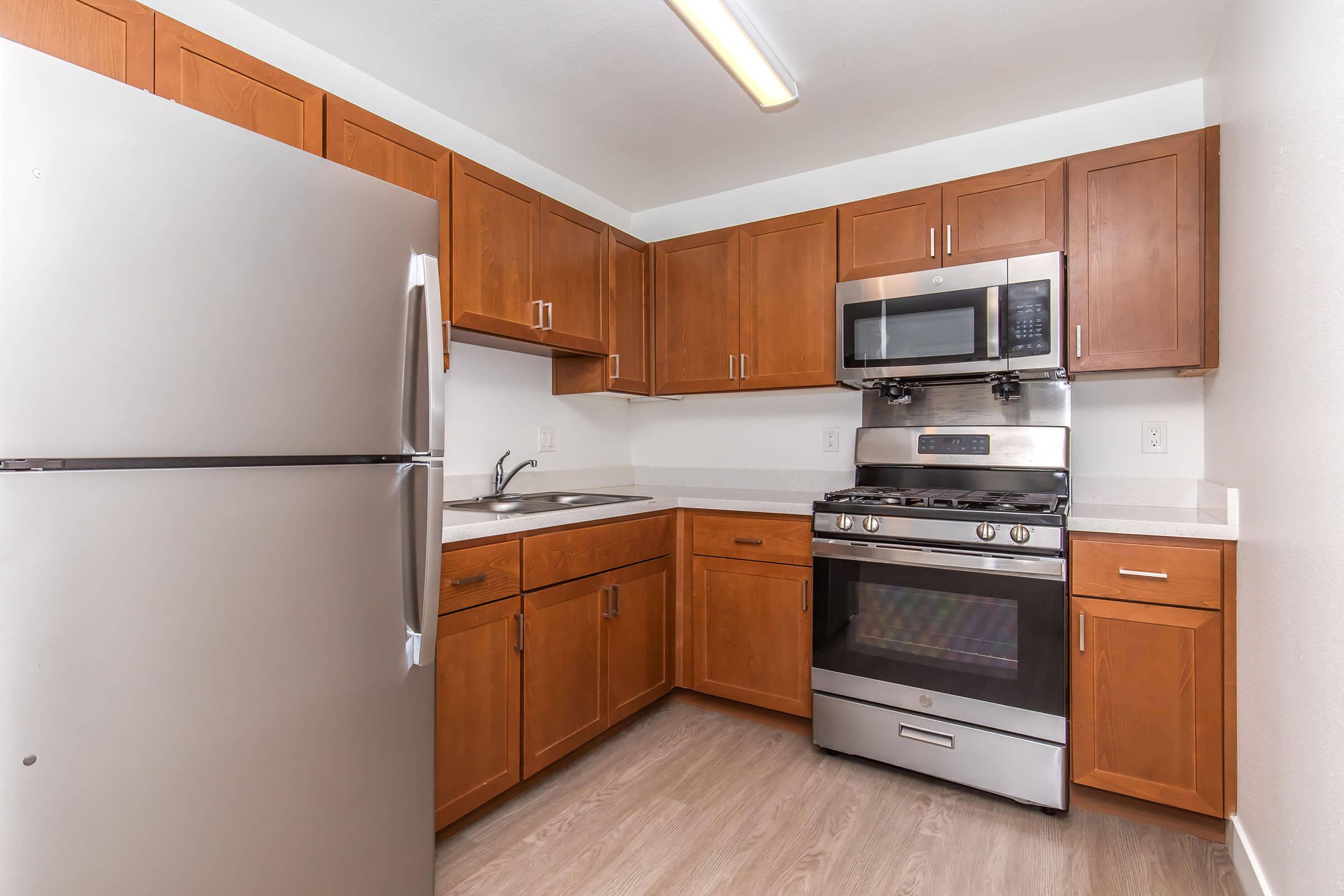 A modern kitchen with wooden cabinets, a stainless steel refrigerator, an oven range with a microwave above, and a sink. The countertop is light-colored, complementing the cabinetry. The room has a bright, clean appearance with a neutral wall color and a light wooden floor.
