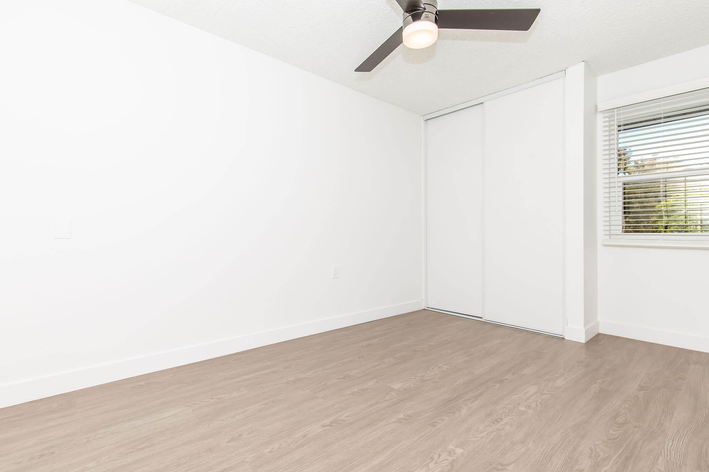 A bright, minimalistic room featuring light-colored walls, a ceiling fan, and a large window with blinds. The floor is covered in light wood laminate, and there is a sliding closet door on one side. The space is empty, creating an open and airy atmosphere.