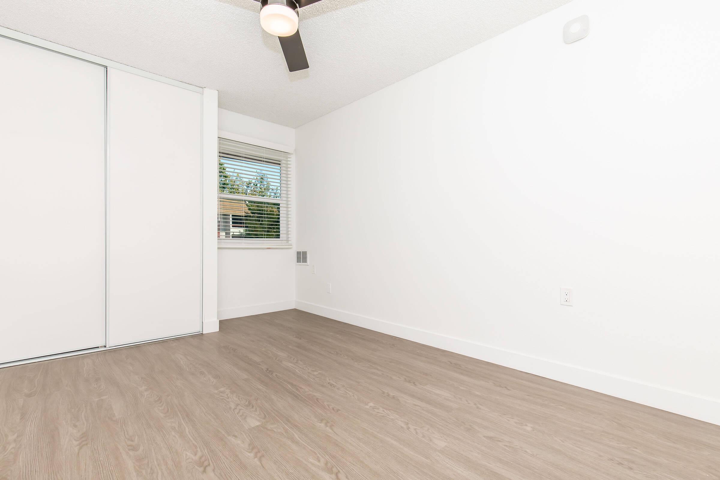 A clean, minimalist room featuring light-colored walls and a ceiling fan. The floor is a medium wood laminate, and there is a window allowing natural light to enter. One wall has sliding doors, and the space appears empty, giving a sense of openness and potential for personalization.