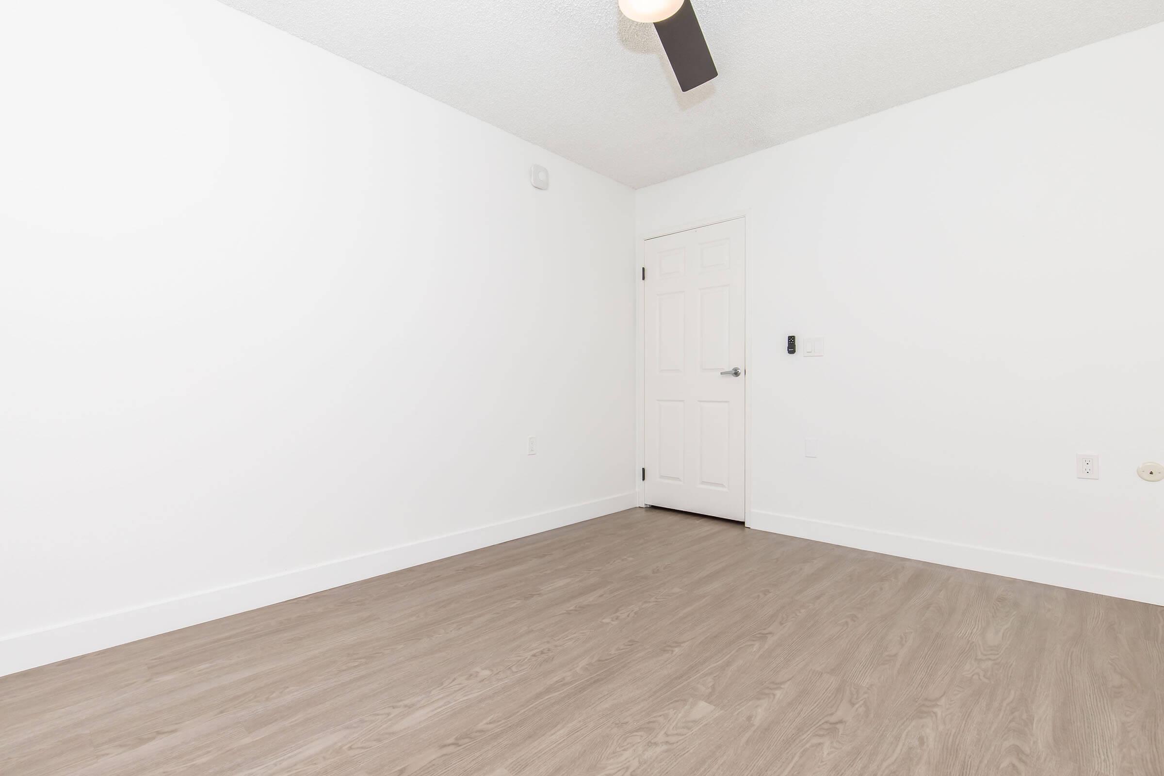A sparsely furnished room with white walls and a light brown wooden floor. A ceiling fan is visible, and there is a closed white door on the right side of the image. The room appears bright and empty, suitable for various uses or as a blank canvas for decoration.