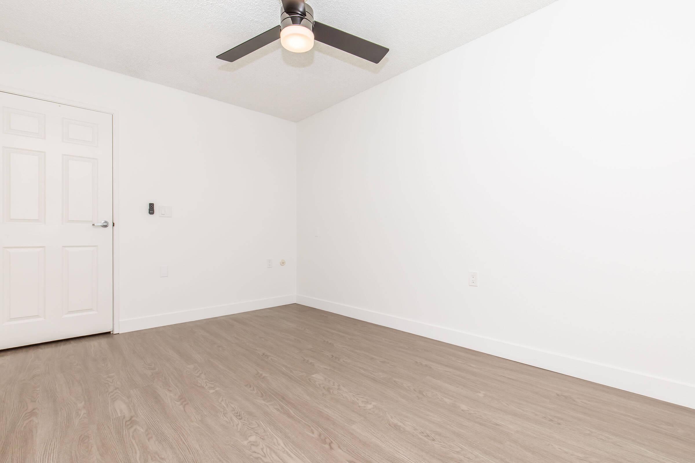 A spacious, empty room with light brown laminate flooring. The walls are painted white and a ceiling fan is installed. There is a closed white door on the left side of the image, and a wall-mounted device visible near the door. The room has a bright, minimalist aesthetic.