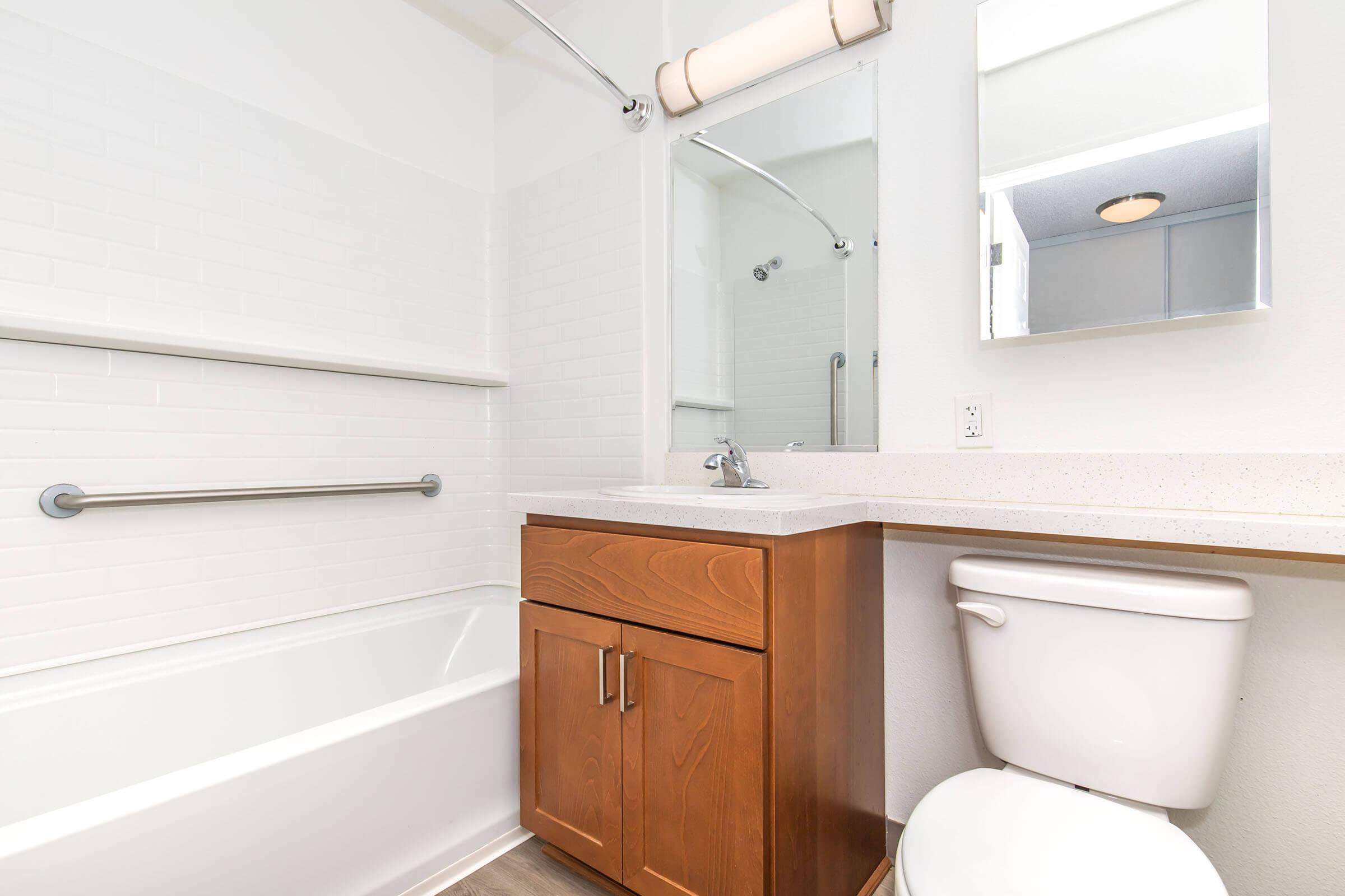 A clean, modern bathroom featuring a white tile shower with a bathtub, a small wooden vanity with a sink, a mirror, and a toilet. Soft lighting from a ceiling fixture illuminates the space, highlighting the simple, functional design. The overall decor is bright and minimalist.