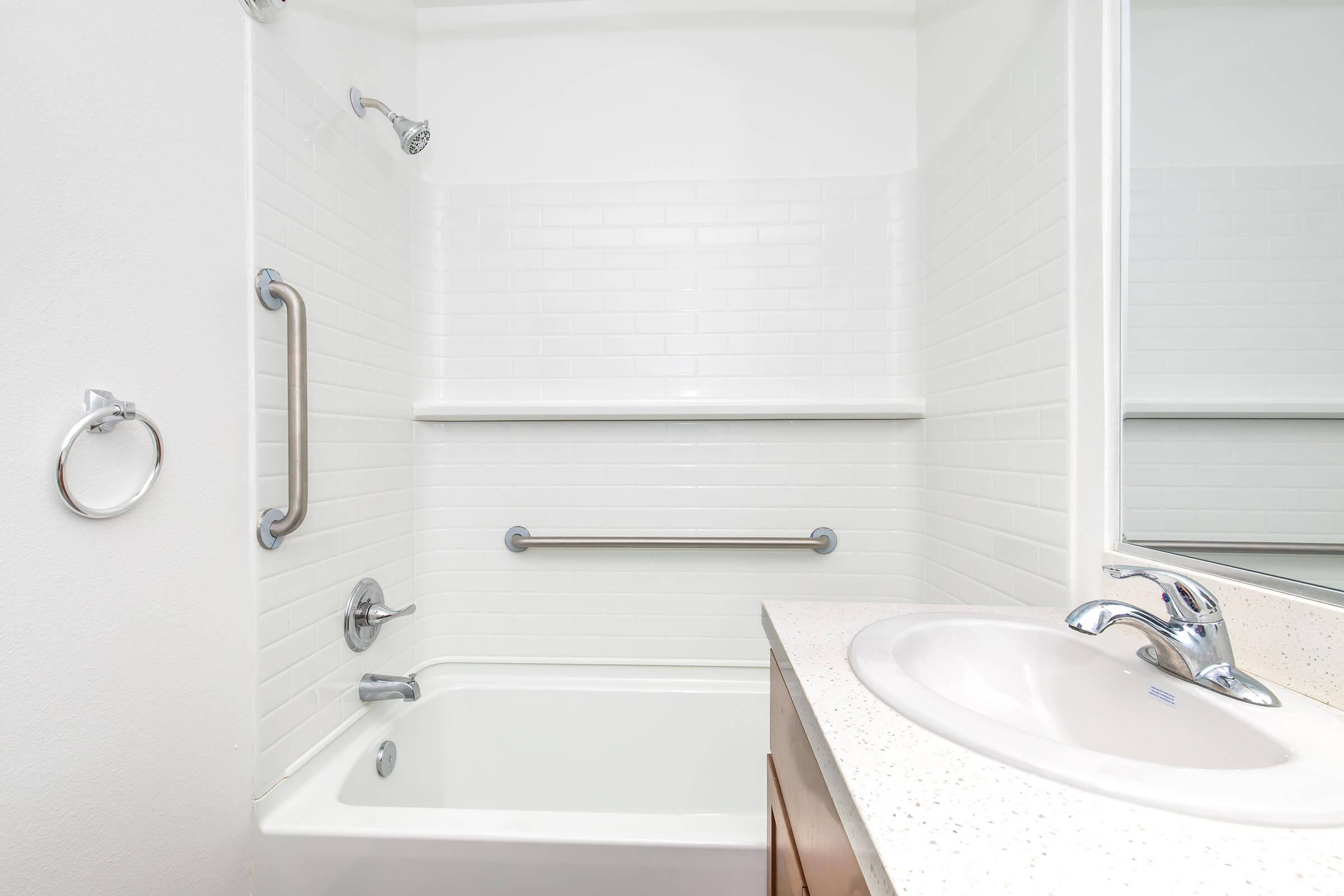 A clean and modern bathroom featuring a bathtub with a shower, white tiled walls, grab bars for safety, and a sink with a single faucet. The room has a minimalistic design with a bright, uncluttered appearance. A mirror is reflected above the sink area.