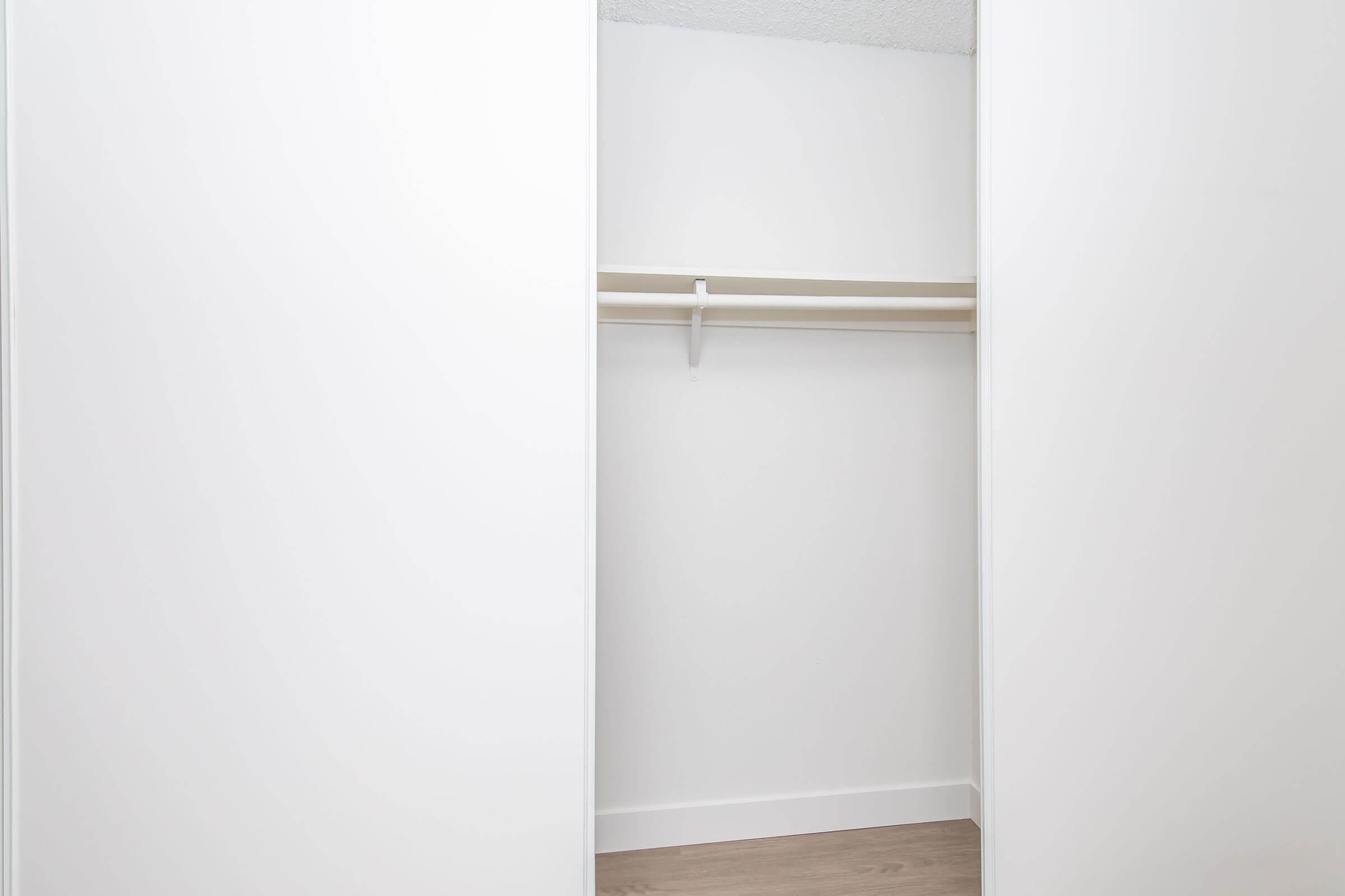 An empty closet featuring white walls and a wooden floor. The closet has sliding white doors and a single shelf inside, with no items or additional furnishings present.