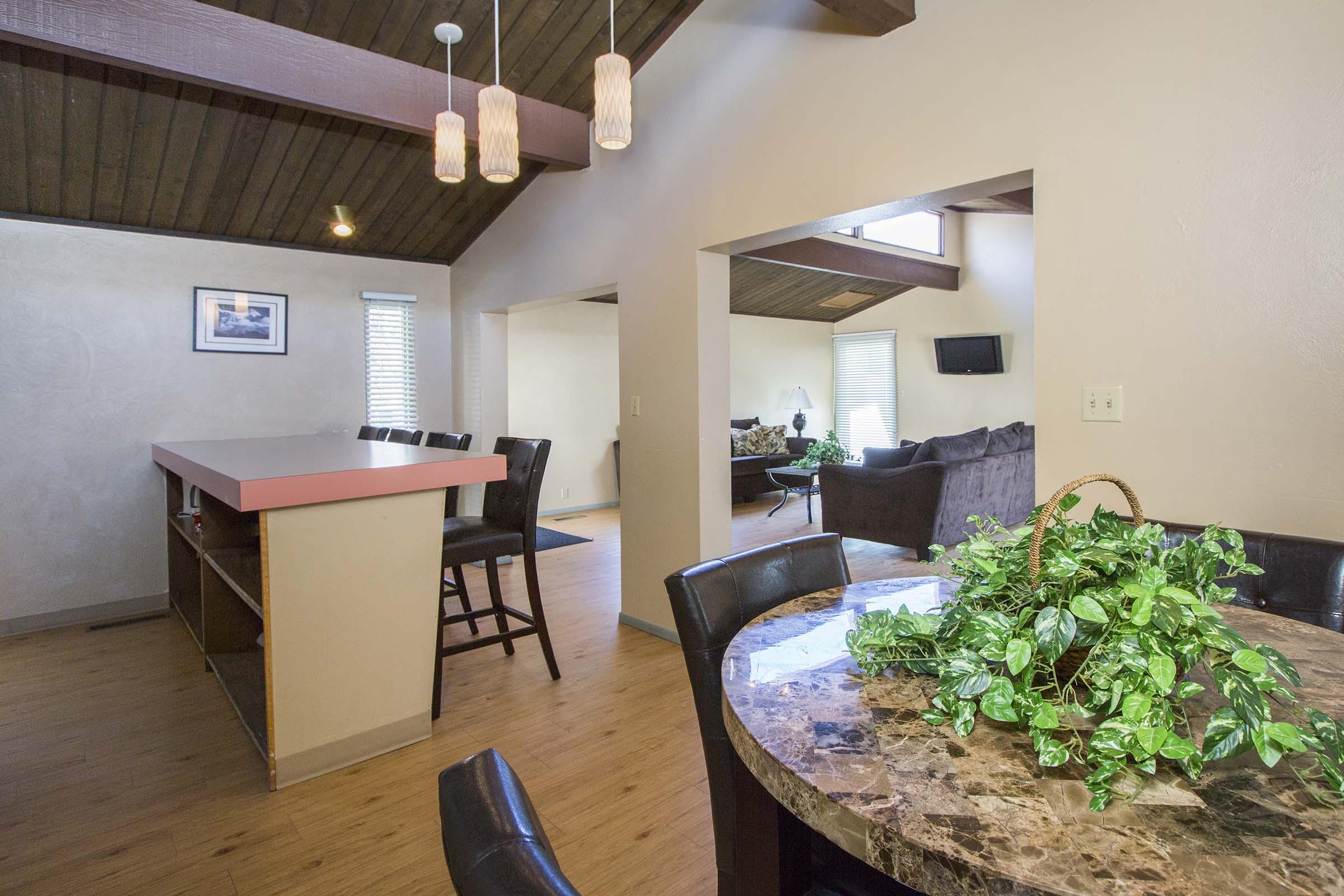 Interior of a modern home with an open layout featuring a kitchen island with bar stools, a dining table with a marble top and a potted plant. The ceiling has wooden beams, and there are bright pendant lights hanging down. In the background, a living area with a sofa and TV is visible.